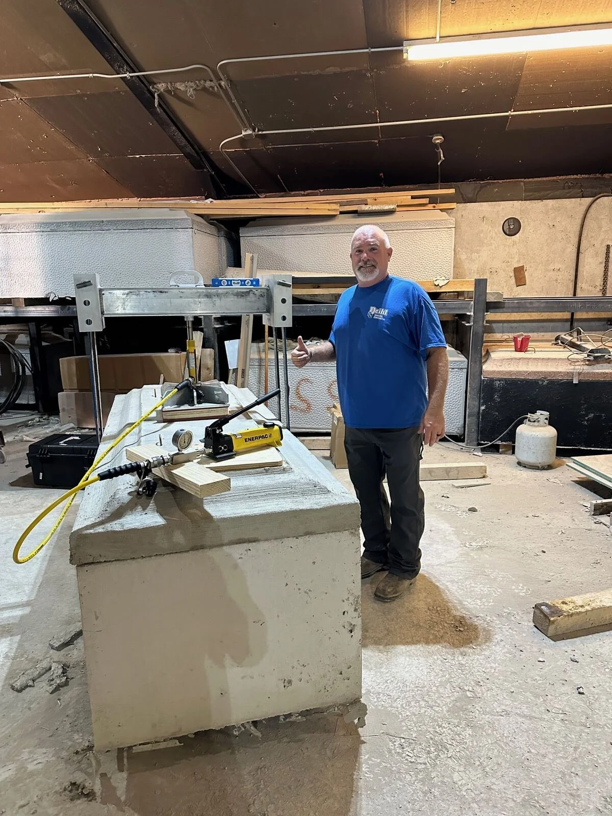 A man standing in a woodworking shop giving a thumbs up, with tools and wood on the workbench, and construction materials and equipment in the background.