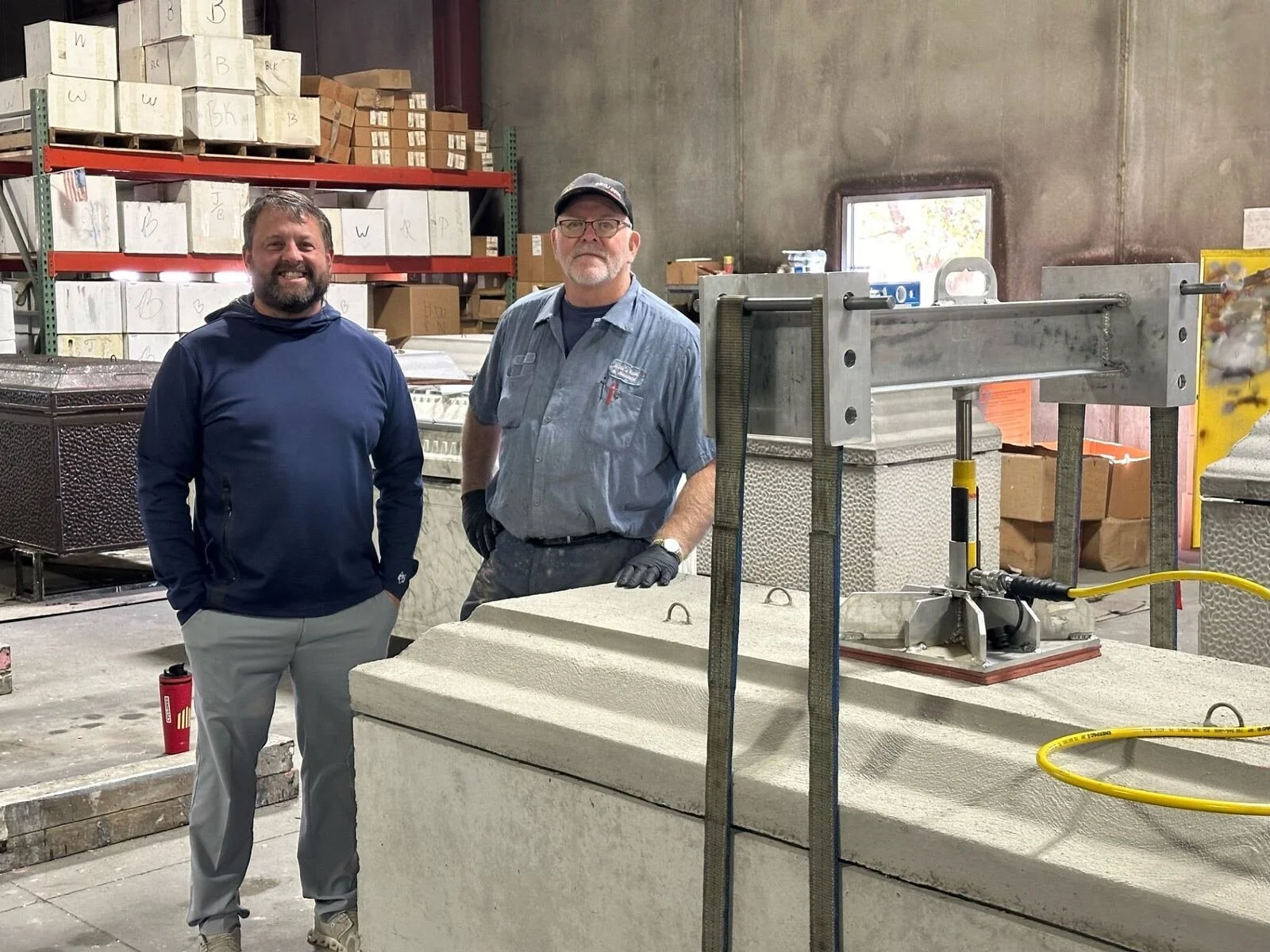 Two men standing in an industrial warehouse surrounded by large stone blocks, metal shelving with boxes in the background, and a machine or tool in front of them. One man is smiling, wearing a navy hoodie and light gray pants. The other is wearing a blue shirt, glasses, and a cap.