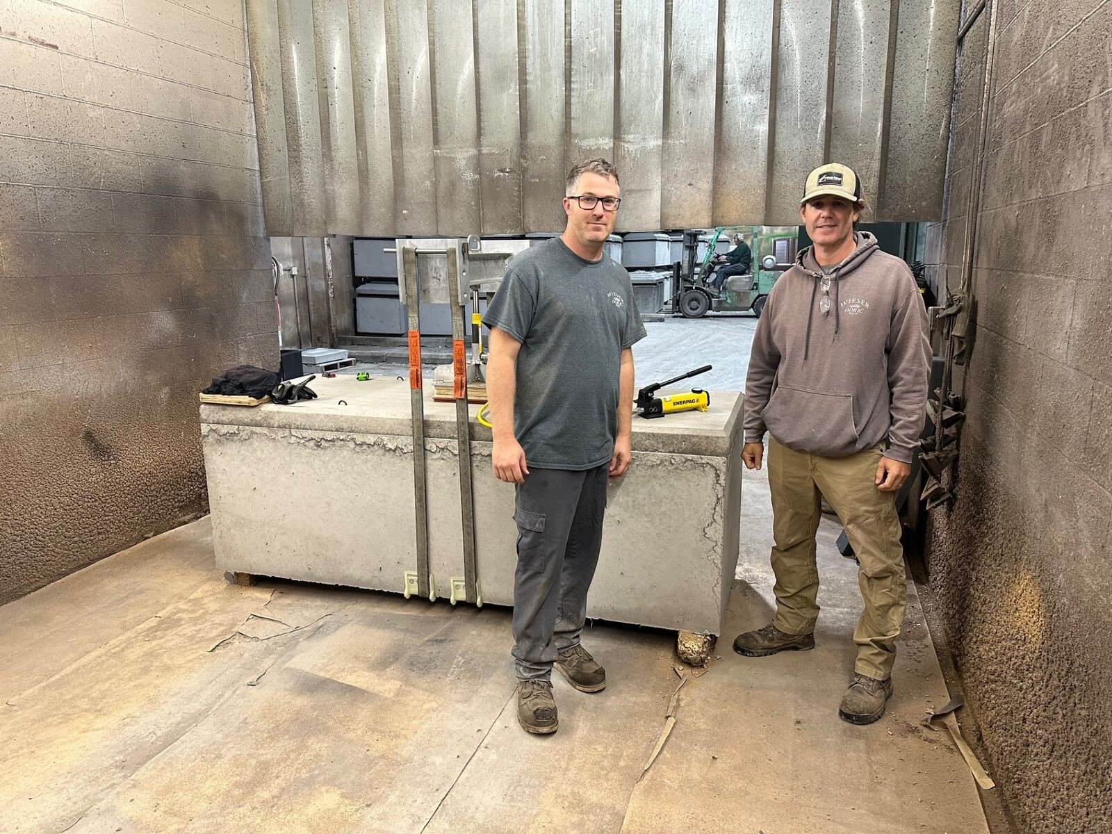 Two workers standing in an industrial setting with concrete walls and floor. One worker is wearing a gray t-shirt and gray pants, and the other is wearing a brown hoodie and tan pants. There are tools and equipment on a large rectangular concrete block in front of them.