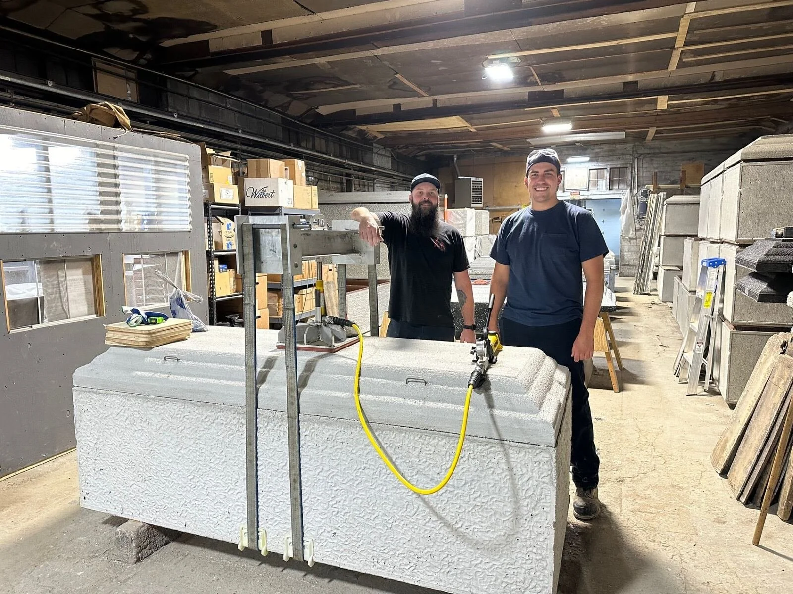 Two men standing behind a large white textured stone block in a workshop, surrounded by construction materials and tools, with boxes and shelves in the background.