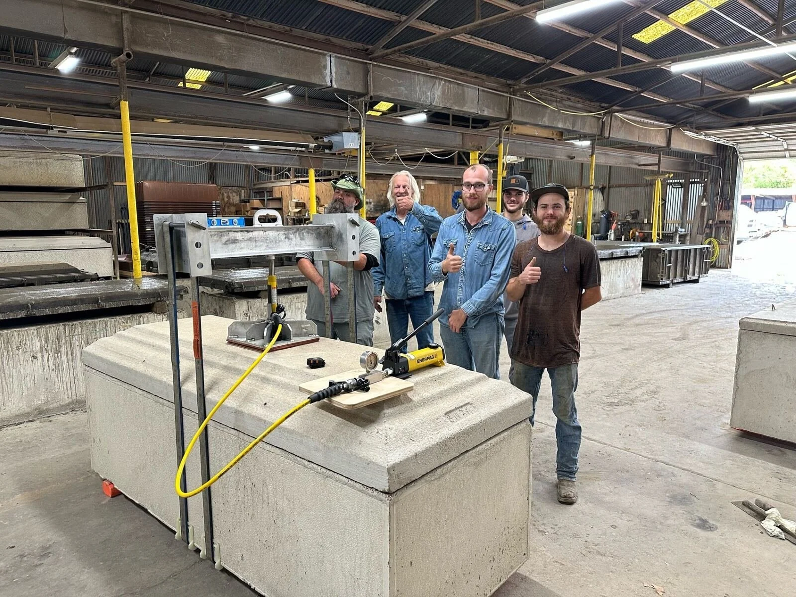 Five men standing inside a workshop or factory. They are smiling and giving thumbs up. There is a large concrete block with equipment on it in the foreground, with a yellow coiled tube attached. The workshop has metal walls, a high ceiling with exposed pipes, and various tools and materials in the background.