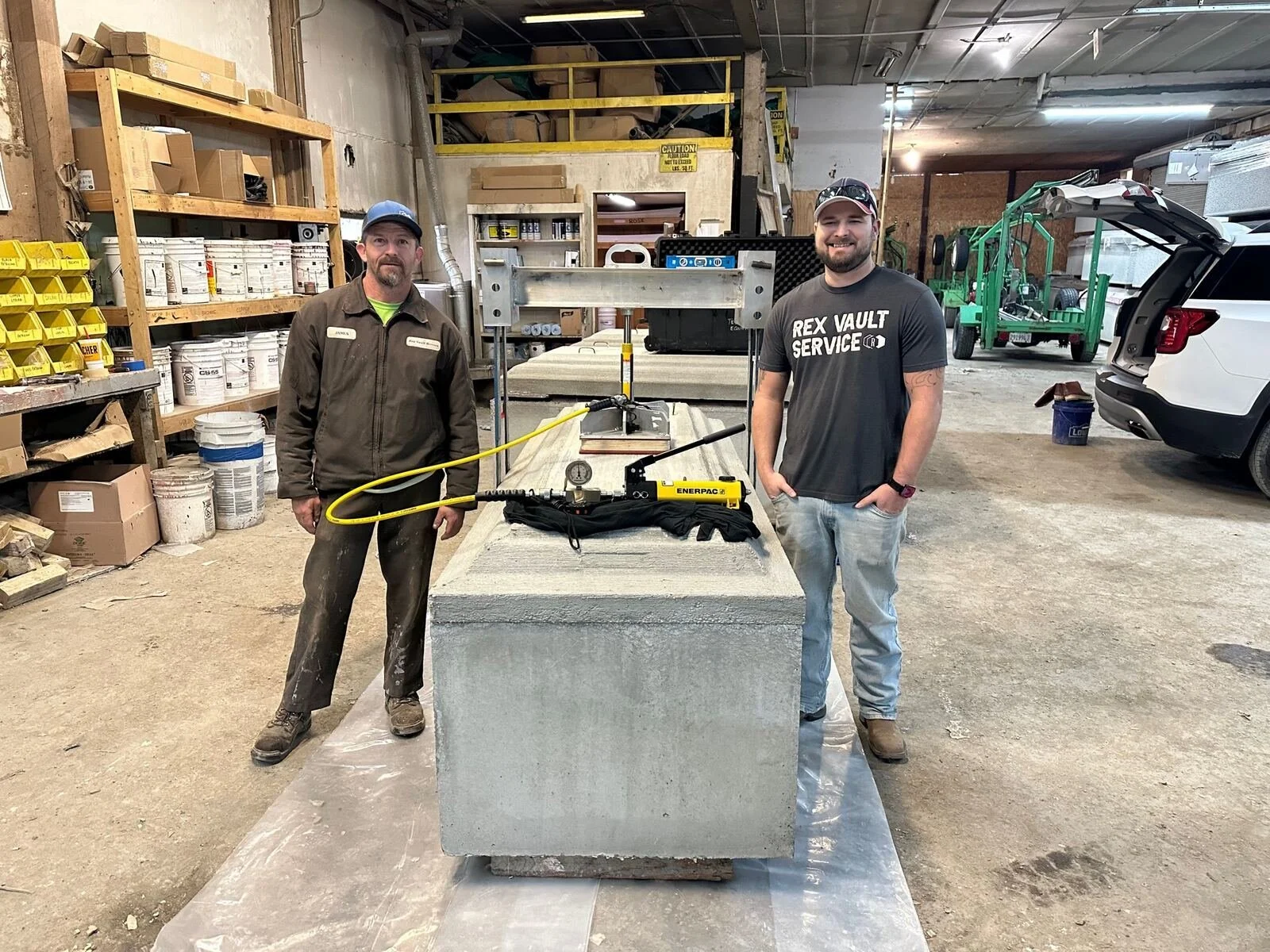 Two men standing on either side of large concrete block, using a hydraulic tool connected by a yellow hose, inside a workshop or warehouse with shelves filled with supplies and tools.