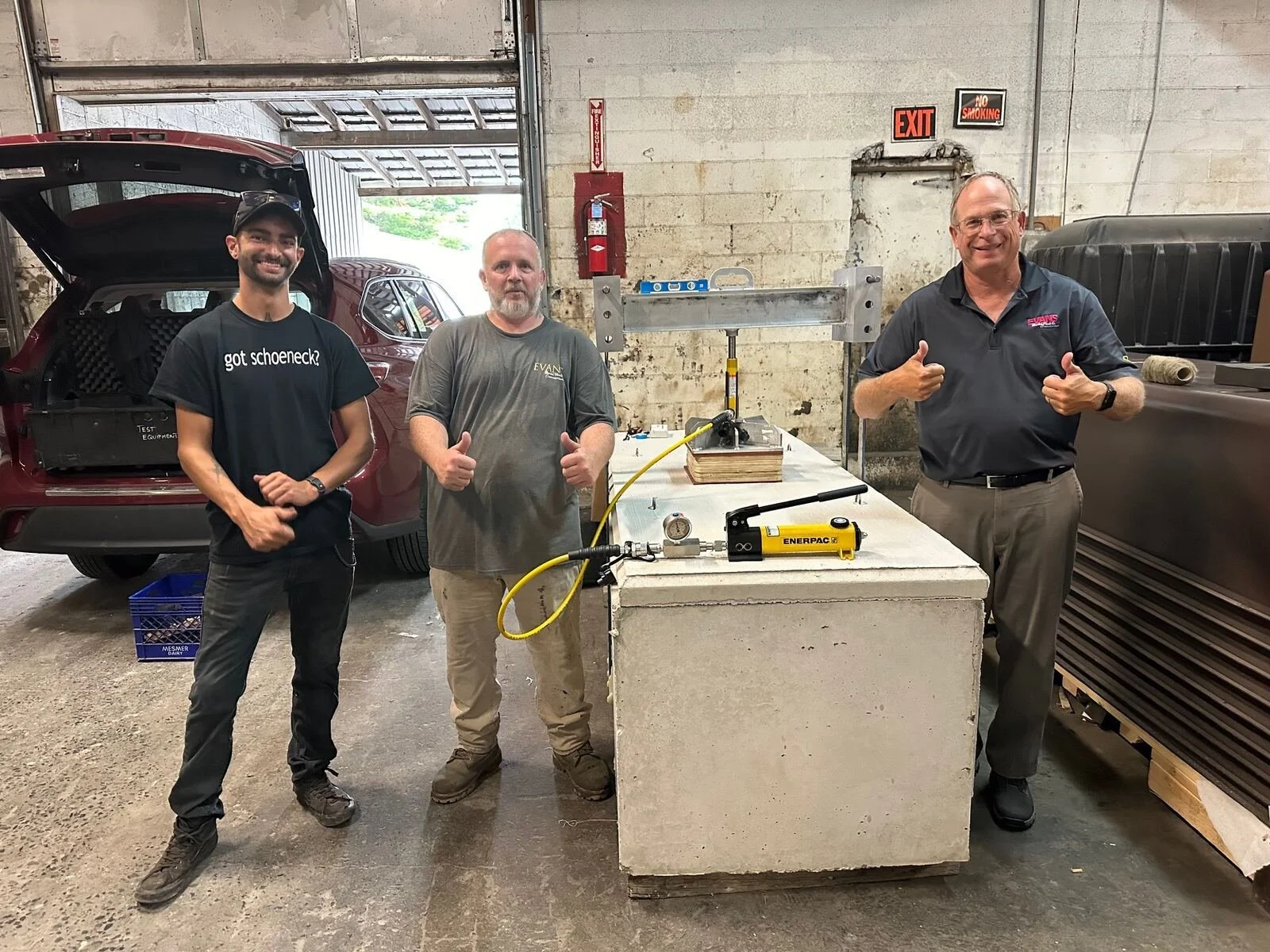Three men standing inside a garage with cars in the background. The man on the left is wearing a black t-shirt and cap, the man in the middle is wearing a gray shirt, and the man on the right is wearing a dark polo shirt. They are all smiling, and the man on the right is giving a thumbs-up. There are tools and equipment on the table in front of them.