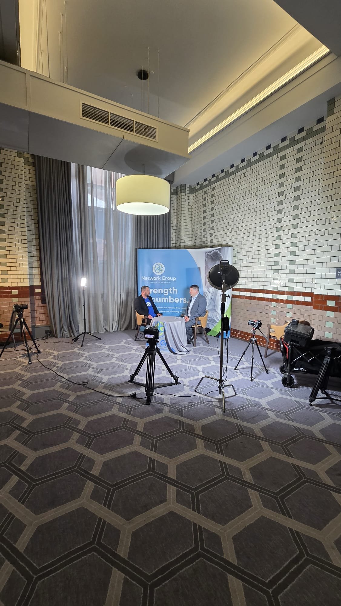 Two men are sitting at a small round table during an interview or discussion, with cameras and lighting equipment set up in a large, well-lit room with patterned carpet, brick walls, and a promotional banner for 'Network Group' behind them.