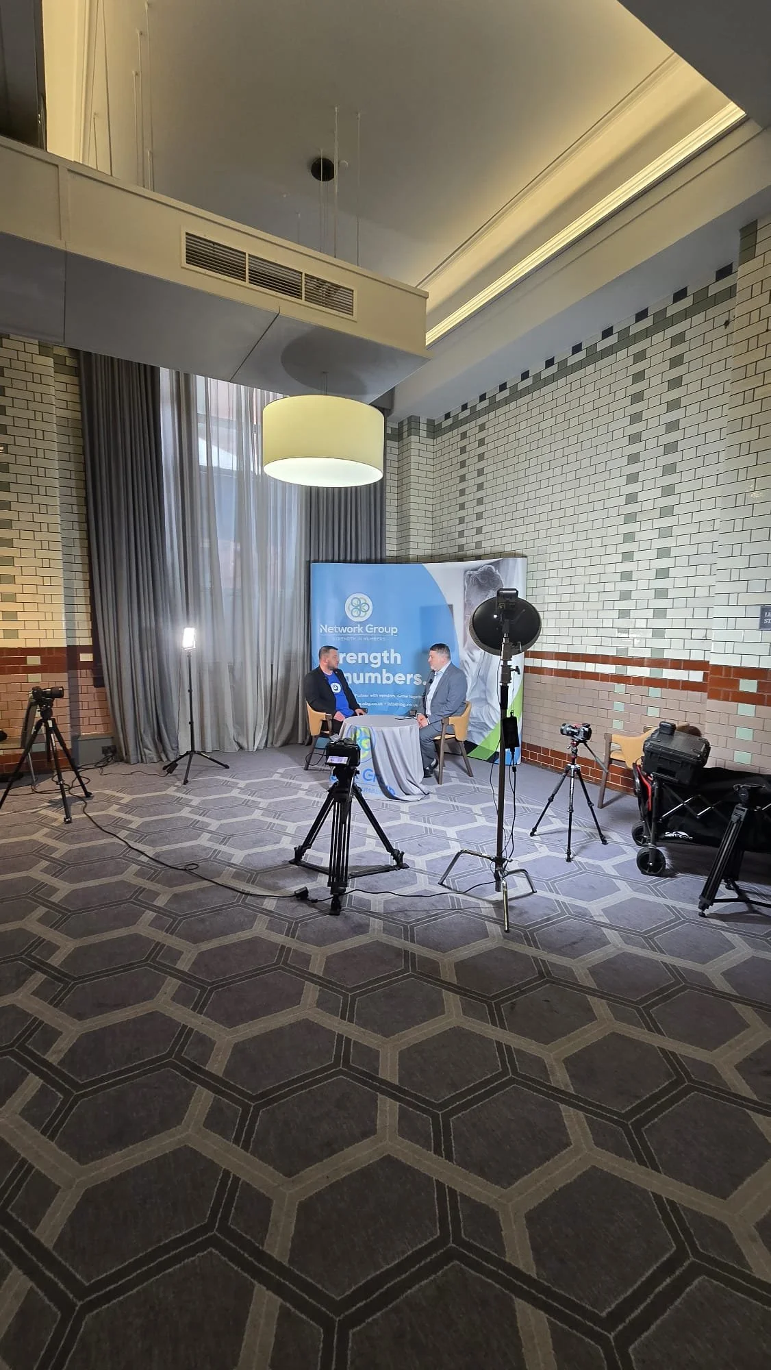 Two men in formal attire sitting across from each other at a small round table during an interview or discussion, with professional camera equipment set up around them, branded backdrop with text, all within a spacious, well-lit conference room with patterned carpet and high ceiling.
