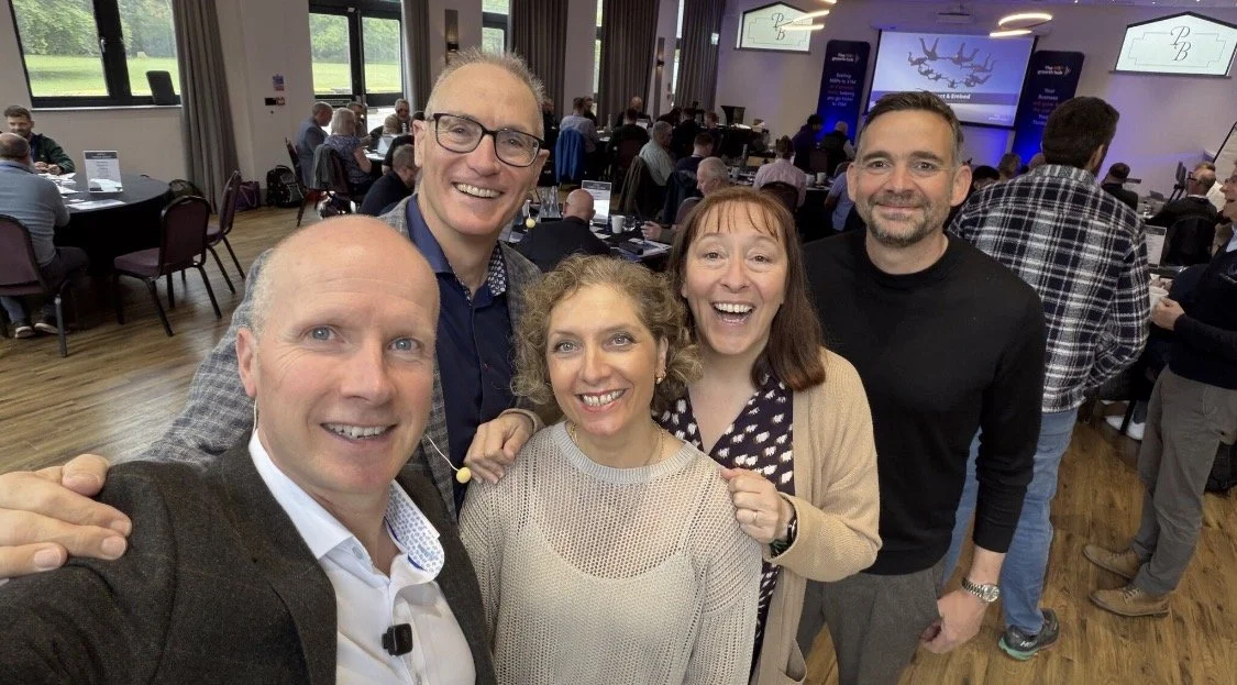 A group of five smiling people taking a selfie at a conference or seminar in a large room with many attendees, tables, and screens.