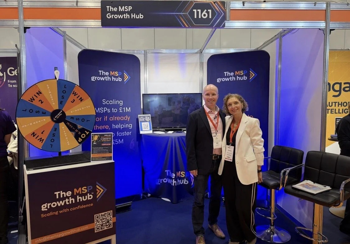 At a trade show booth for The MSP Growth Hub, with two smiling people standing in front, a spinning prize wheel, a TV screen, and banners promoting scaling MSPs to £1M, with branding and technology displays.