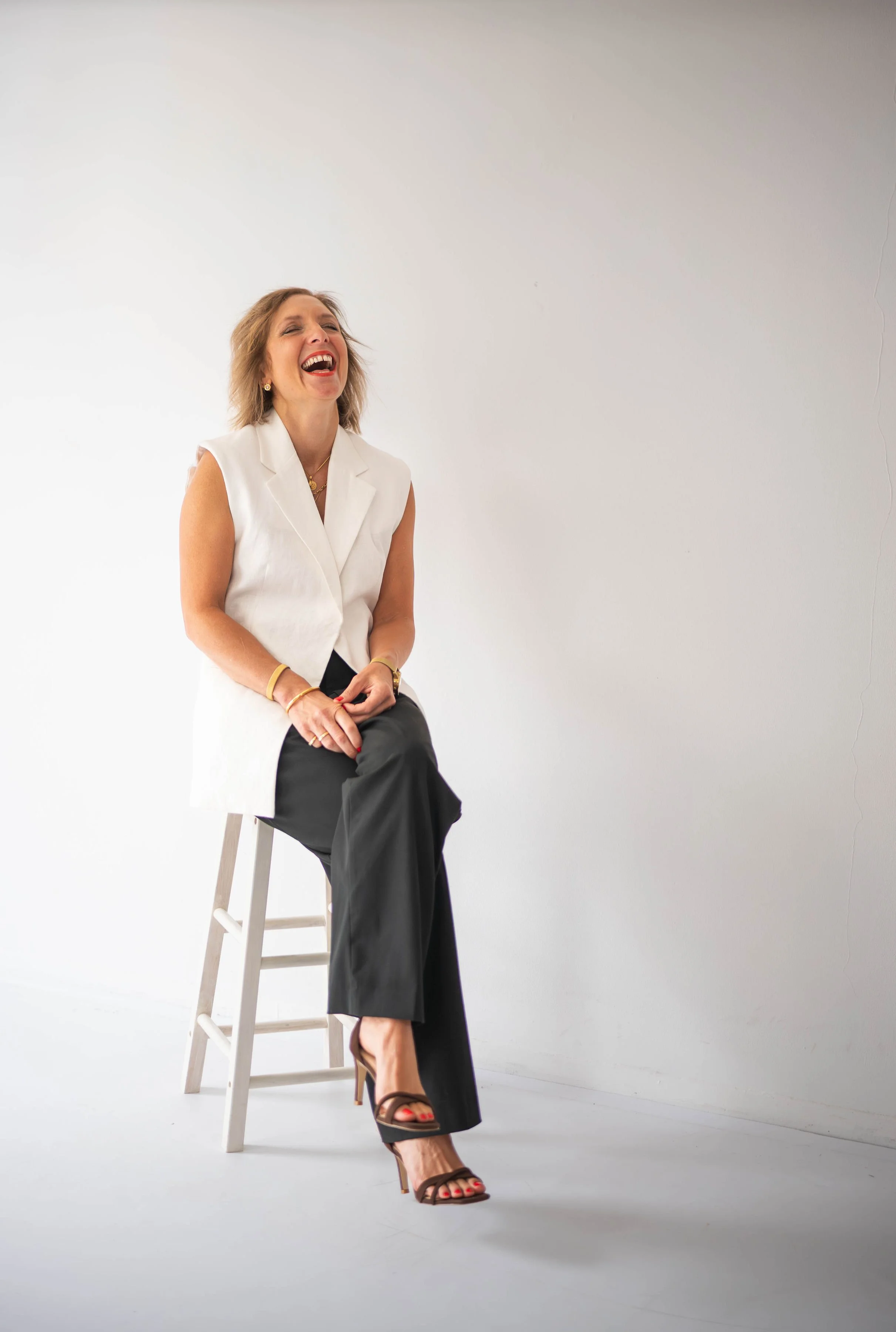 A woman sitting on a stool, laughing with her head tilted back, in front of a plain white background.