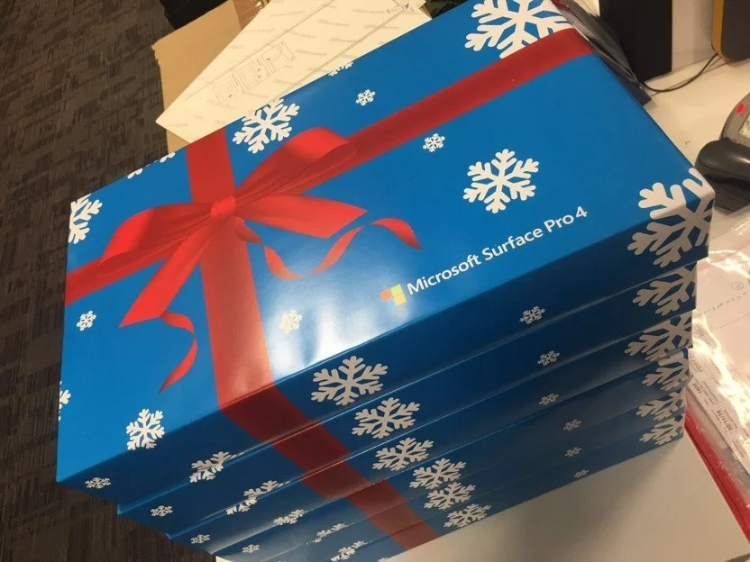 Stack of Microsoft Surface Pro 4 boxes wrapped as Christmas presents with a red bow design, snowflake pattern, and holiday themed design.
