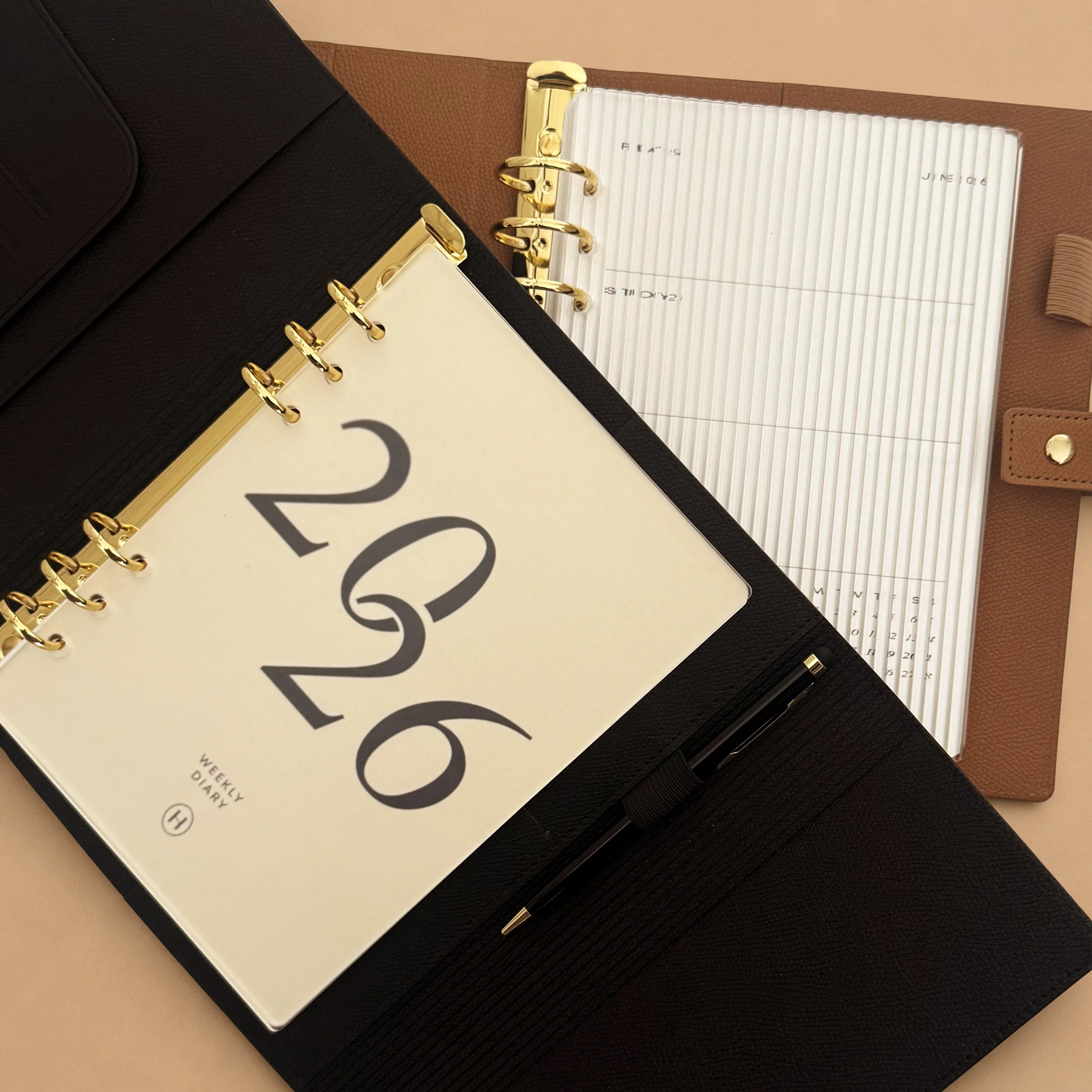 A black planner with gold rings and a cream-colored page inside resting on a black surface. There is a white notebook or planner with gold rings and lined pages partially visible underneath. A pen is lying across the bottom right corner of the black planner.
