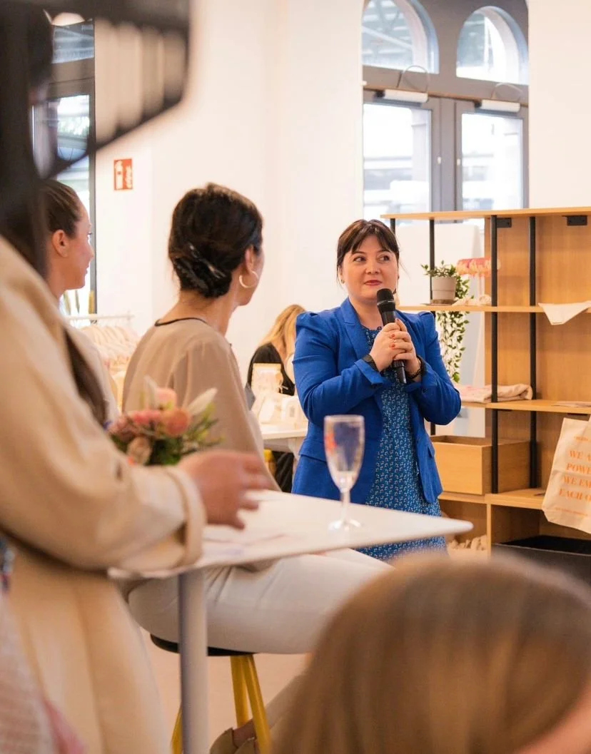 Lela Griessbach in einem blauen Blazer spricht in einem Raum mit mehreren Menschen, die eine Präsentation oder Ansprache verfolgen, während sie ein Mikrofon hält.