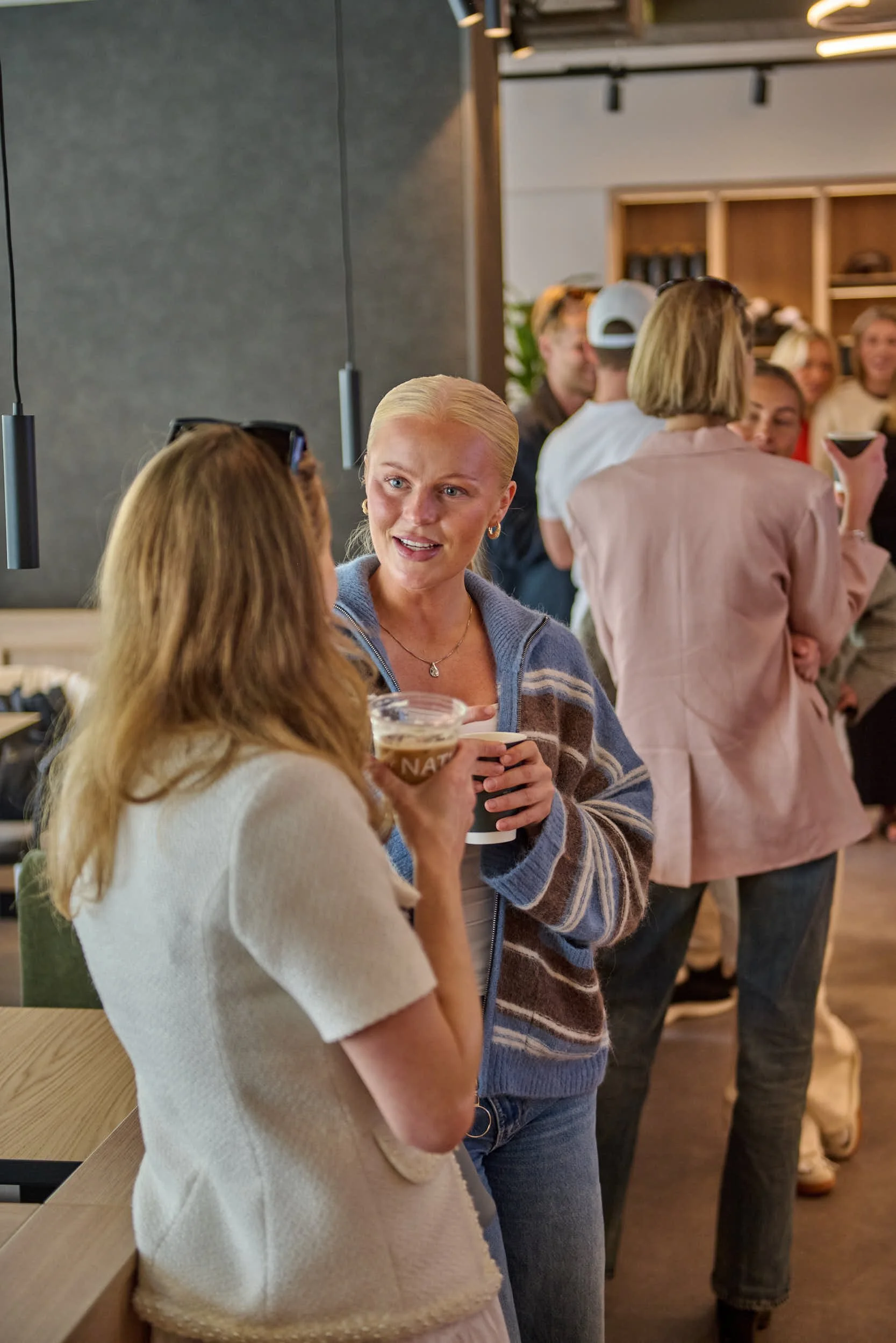 Mennesker snakker og drikker i en sosial setting, sannsynligvis på en bar eller kafé, med flere personer i bakgrunnen.