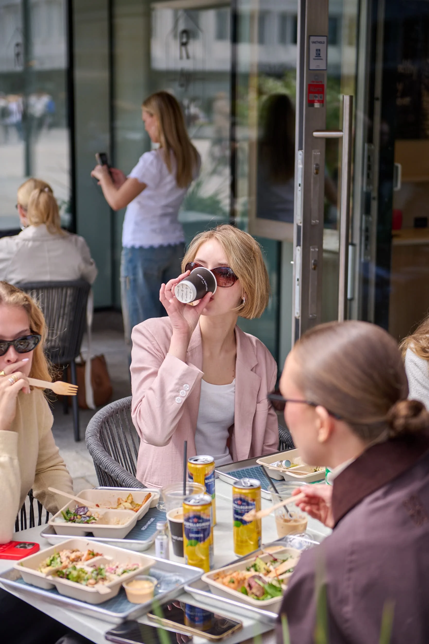 En gruppe mennesker, hovedsakelig kvinner, som spiser ute på et koselig restaurantbord med ulike retter og drikke, mens andre personer i bakgrunnen er opptatt med å bruke telefoner.