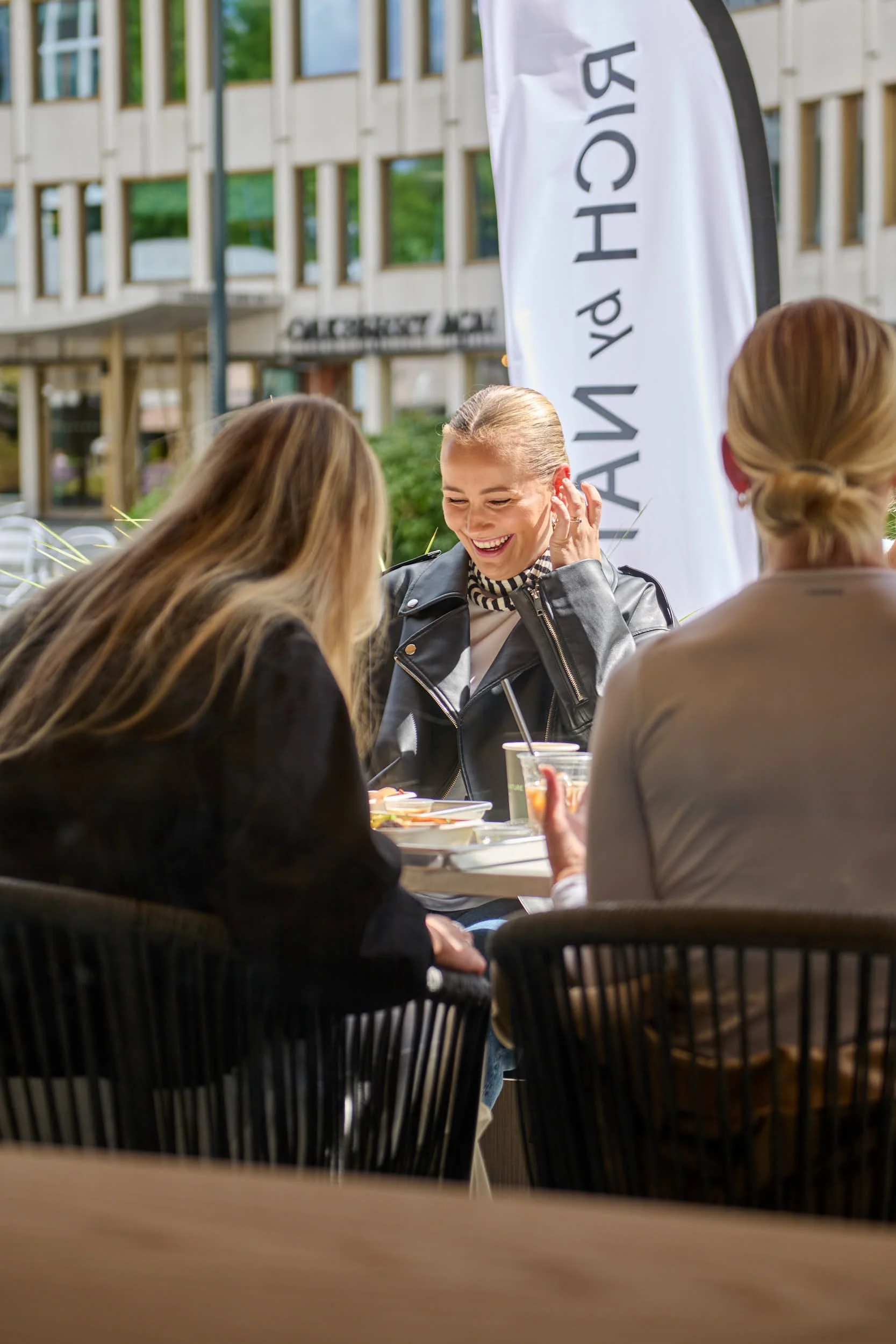 Grupp mennesker som smiler og prater på utendørs kafé med en hvit banner som har svart tekst, i bakgrunnen et moderne bygg med store vinduer.