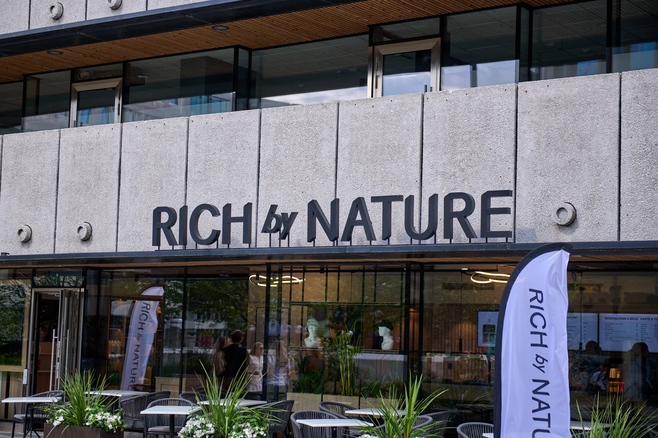 Utsiden av en moderne bygning med store glassvinduer, skilt med teksten RICH BY NATURE, utendørs sitteplasser, blomster og vimpler foran inngangen.