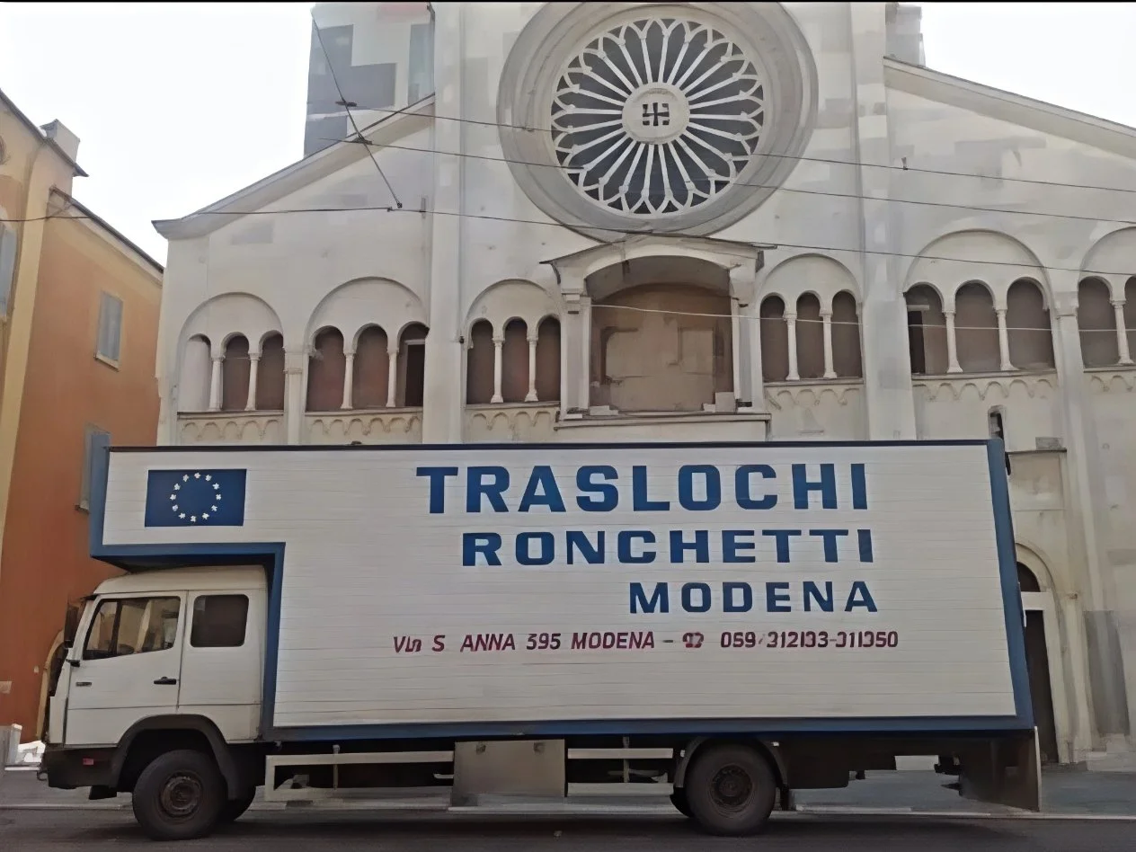 Un camion bianco con una scatola gigante sulla parte laterale con scritto 'Traslochi Ronchetti Modena', parcheggiato davanti a una chiesa con grandi finestre rotonde e una grande rosa in vetro sopra l'ingresso.