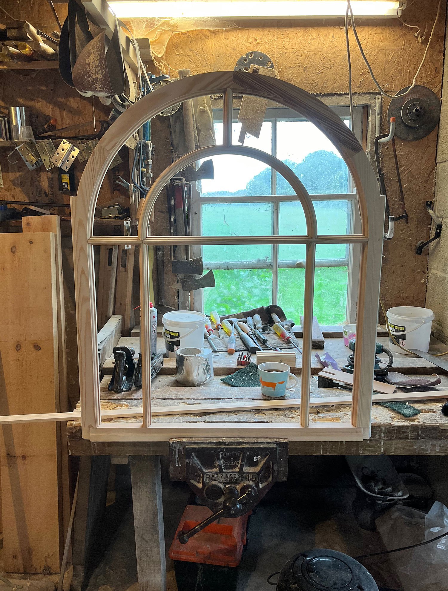 The image shows a workshop with a large unfinished wooden window frame in the center. The workbench is cluttered with various tools, paint cans, and supplies. Behind the window frame, there is a window with a view of green grass and trees outside. The workshop has a rough, unfinished wall with some tools hanging on it and a large window allowing natural light to enter.