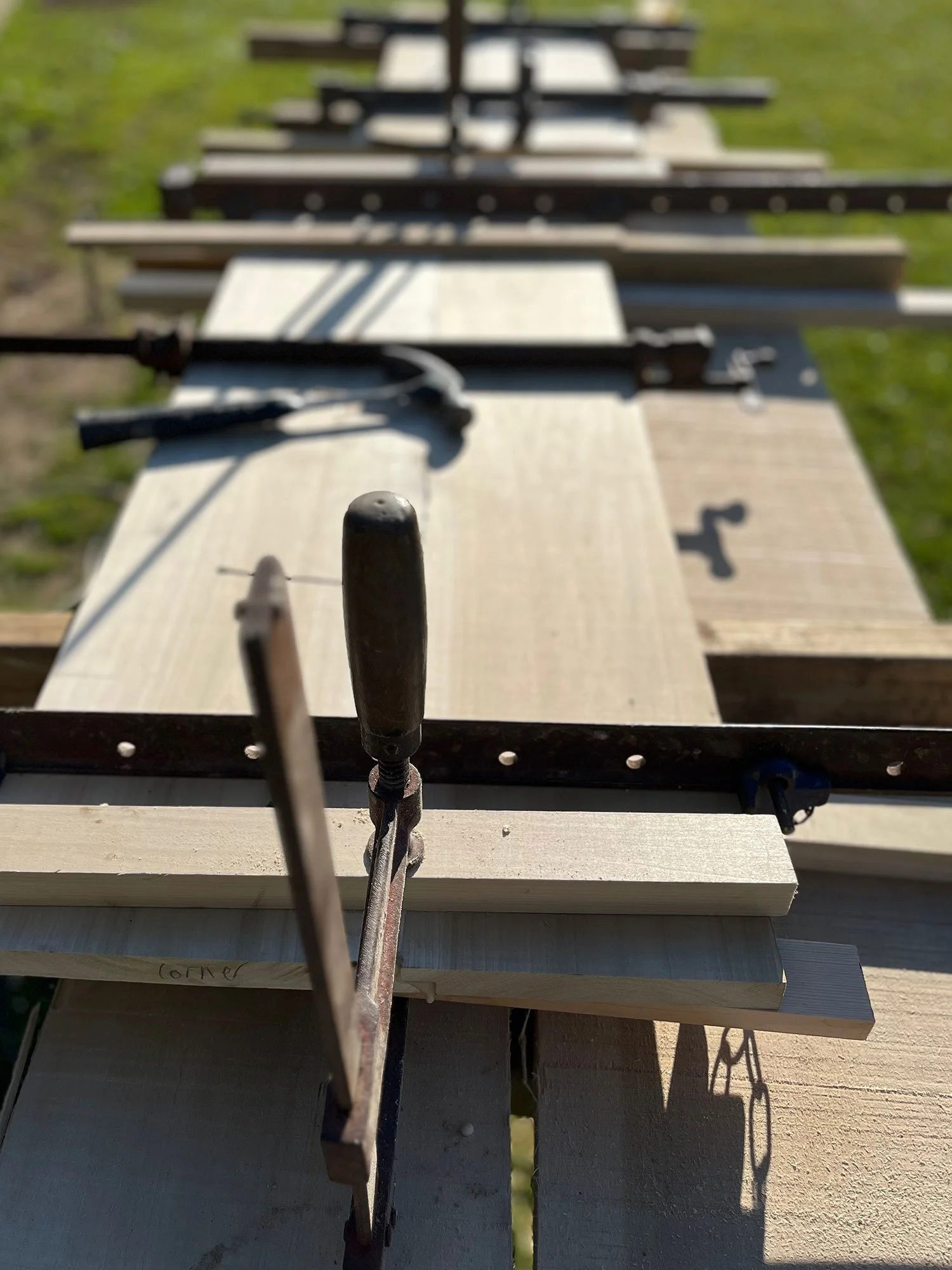 Close-up view of a woodworking clamp tightening on wooden boards outdoors, with grassy area and sunlight in the background.