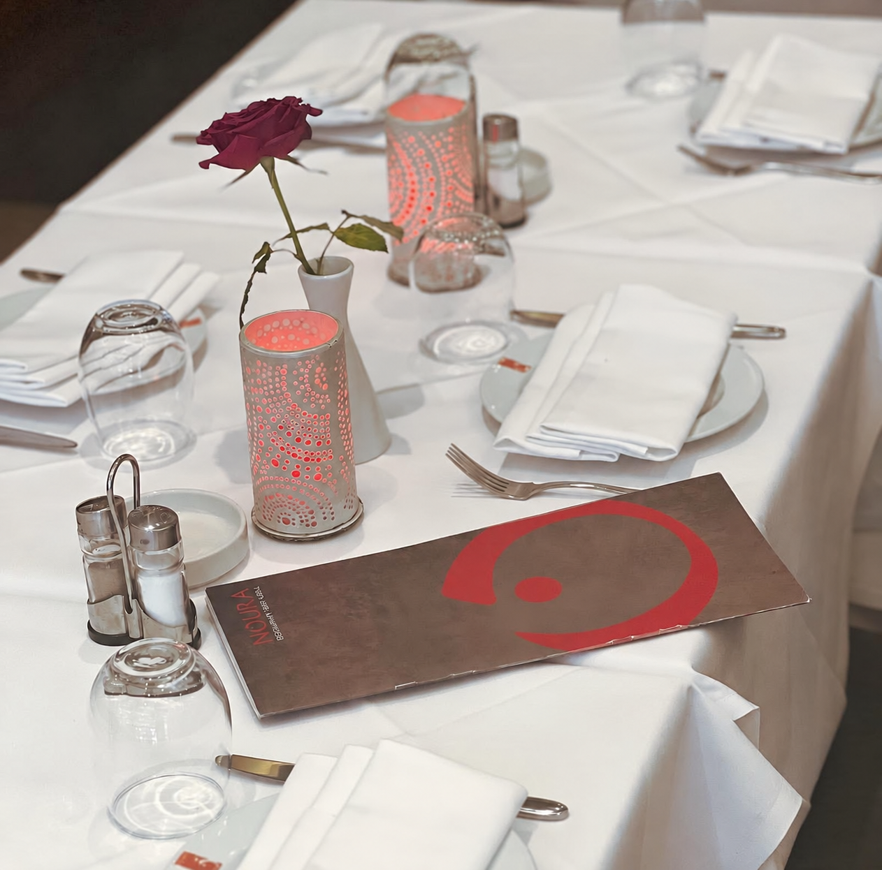 A formal dining table set with white tablecloth, white napkins, and silverware. There are wine glasses turned upside down, a small vase with a red rose, and decorative candles with perforated metal covers. A menu card featuring a large red question mark and the word 'NūRIA' is also on the table.