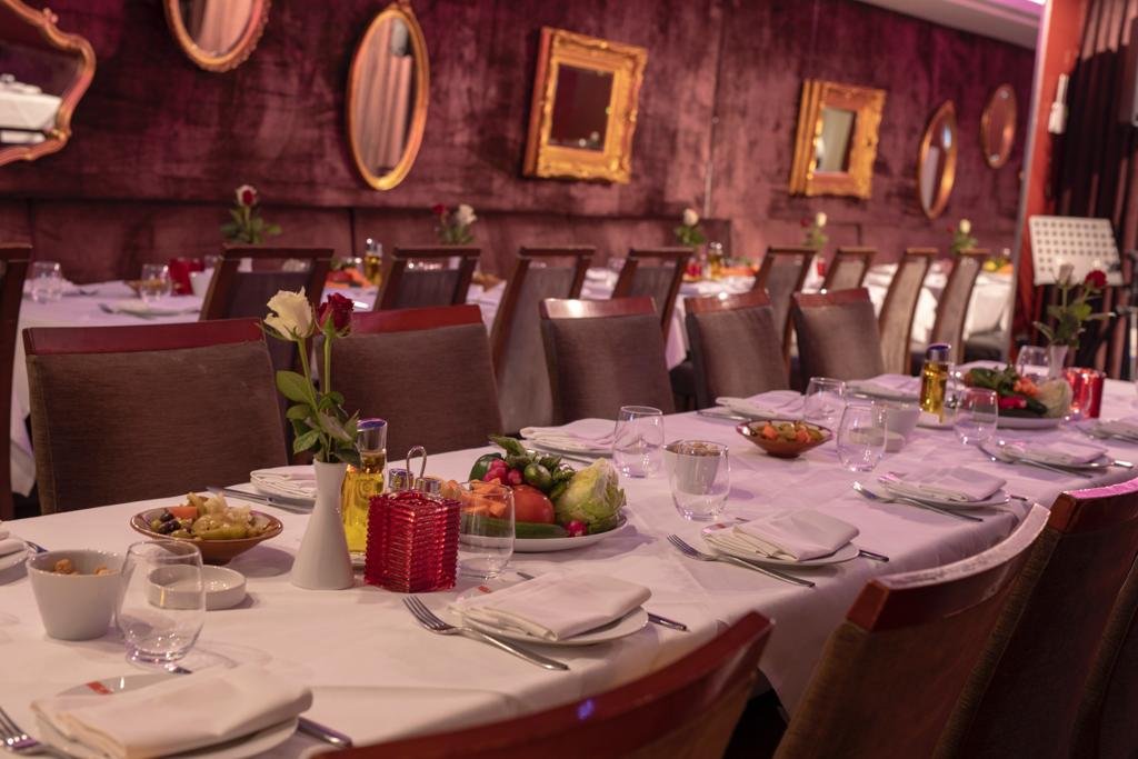 A long dining table set for a meal in a restaurant with many chairs, floral centerpieces, and fruit platters, against a textured red wall decorated with mirrors and frames.