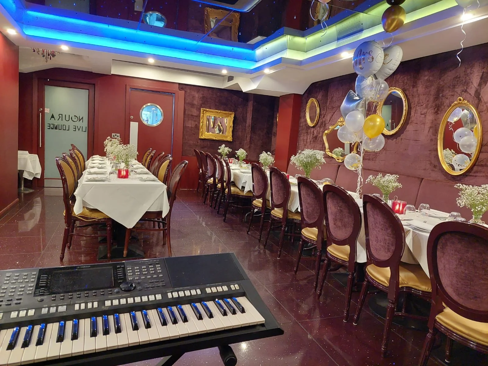 A decorated banquet hall with tables set for a celebration, featuring white tablecloths, floral centerpieces, balloons, and gold-framed mirrors on purple walls.