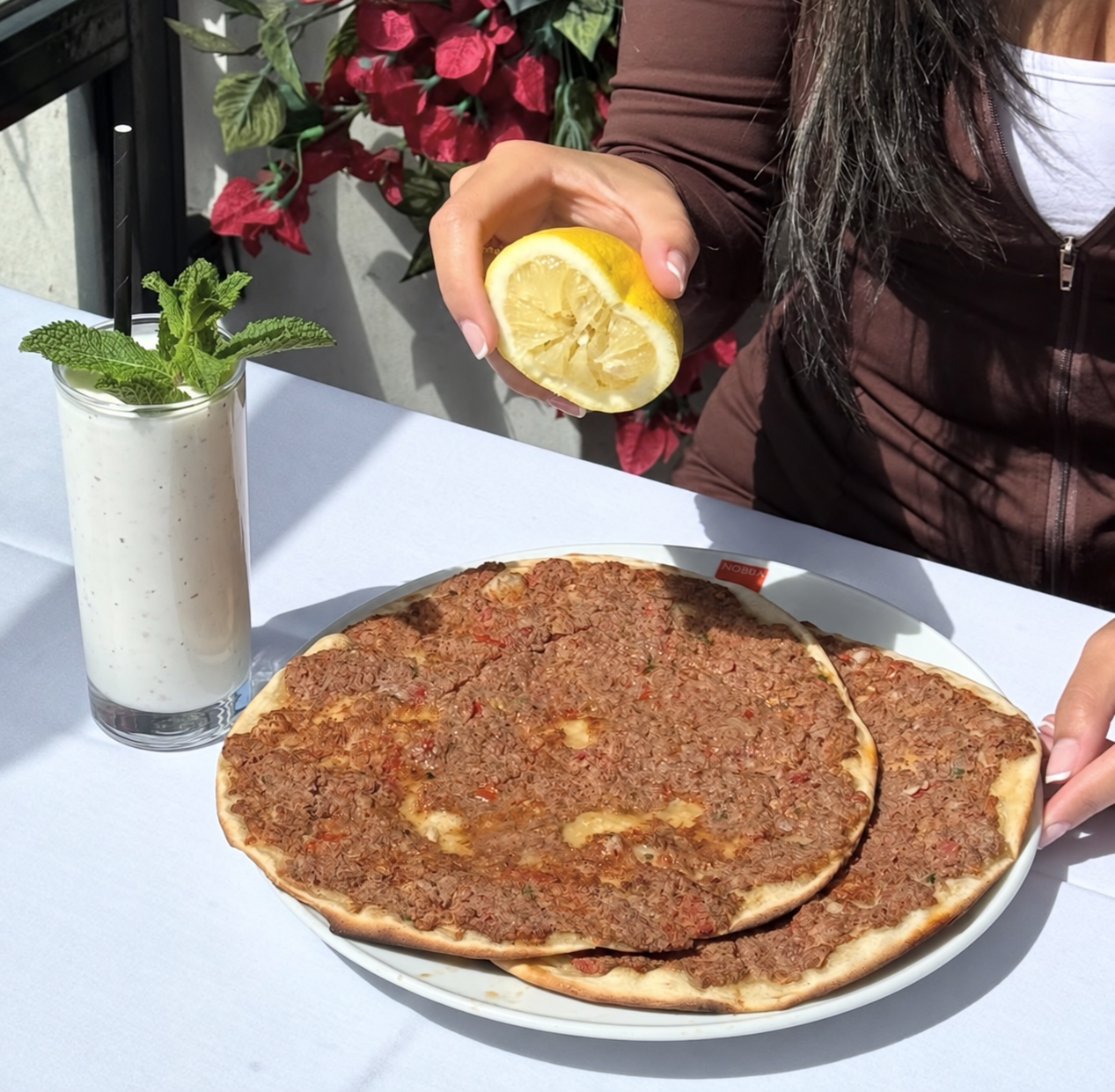 Person squeezing lemon over a plate of traditional Turkish Adana kebab pizza. There is a tall glass of yogurt with mint leaves and a black straw on the table. The person has long dark hair and is wearing a brown jacket and white shirt. Red and green foliage is in the background.