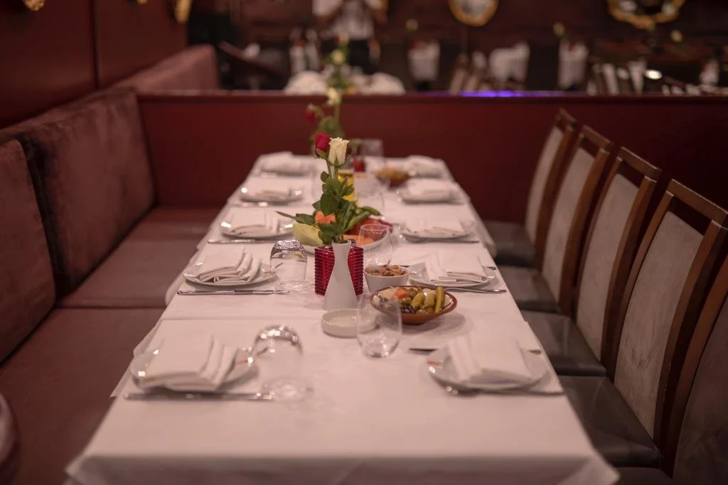 A long dining table set for a meal with white tablecloths, napkins, glasses, and bowls of food. A vase with flowers is placed in the center. The chairs are arranged along both sides of the table in a dimly lit restaurant or banquet room.