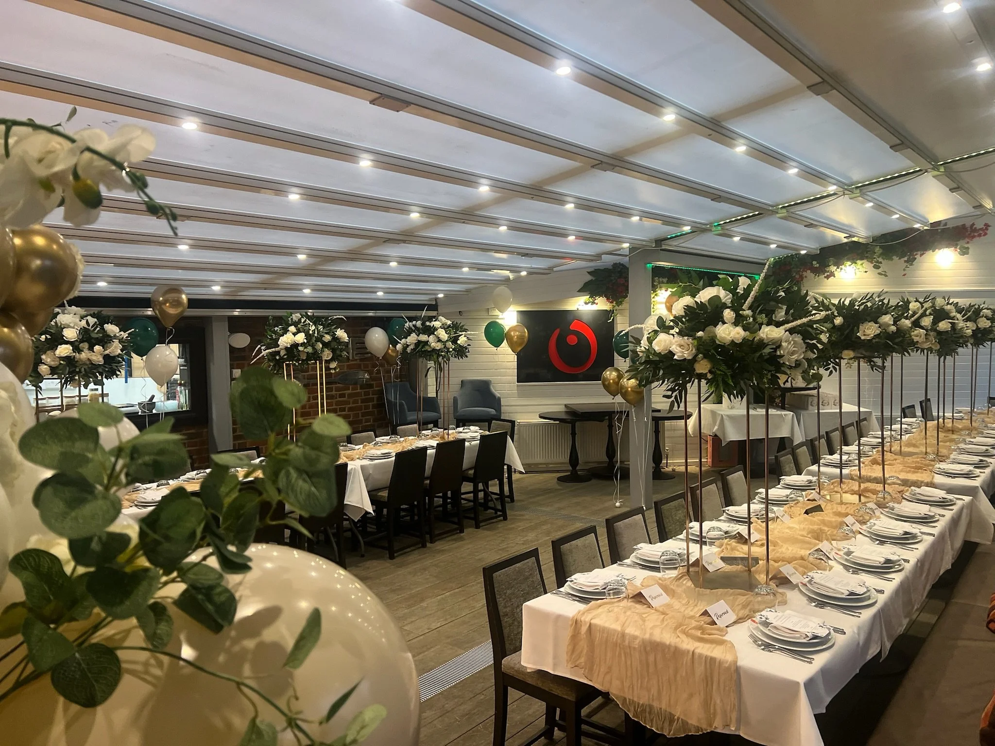 A decorated banquet hall with long tables covered in beige tablecloths, set with plates, silverware, and name cards. The room features floral arrangements with white roses and greenery, gold and white balloons, and ceiling lighting.