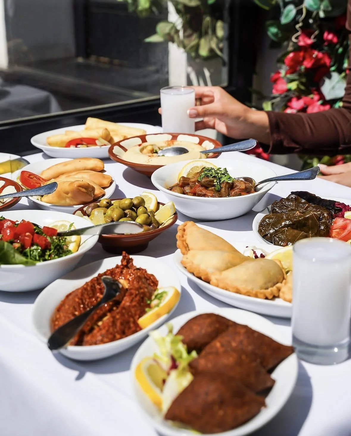 A table filled with various Middle Eastern dishes, including falafel, salads, stuffed grape leaves, hummus, olives, and lamb, with a person holding a glass of water.