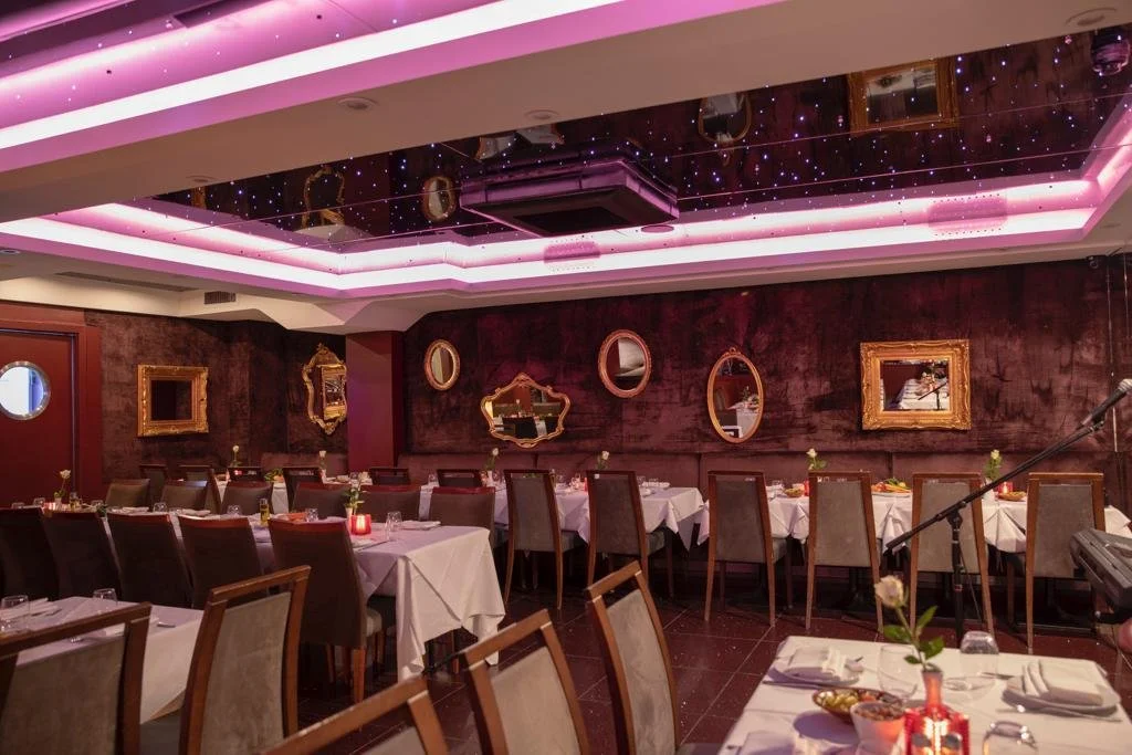 Upscale restaurant interior with dark red velvet walls, gold-framed mirrors, white tablecloths, and chairs, decorated for a special occasion.