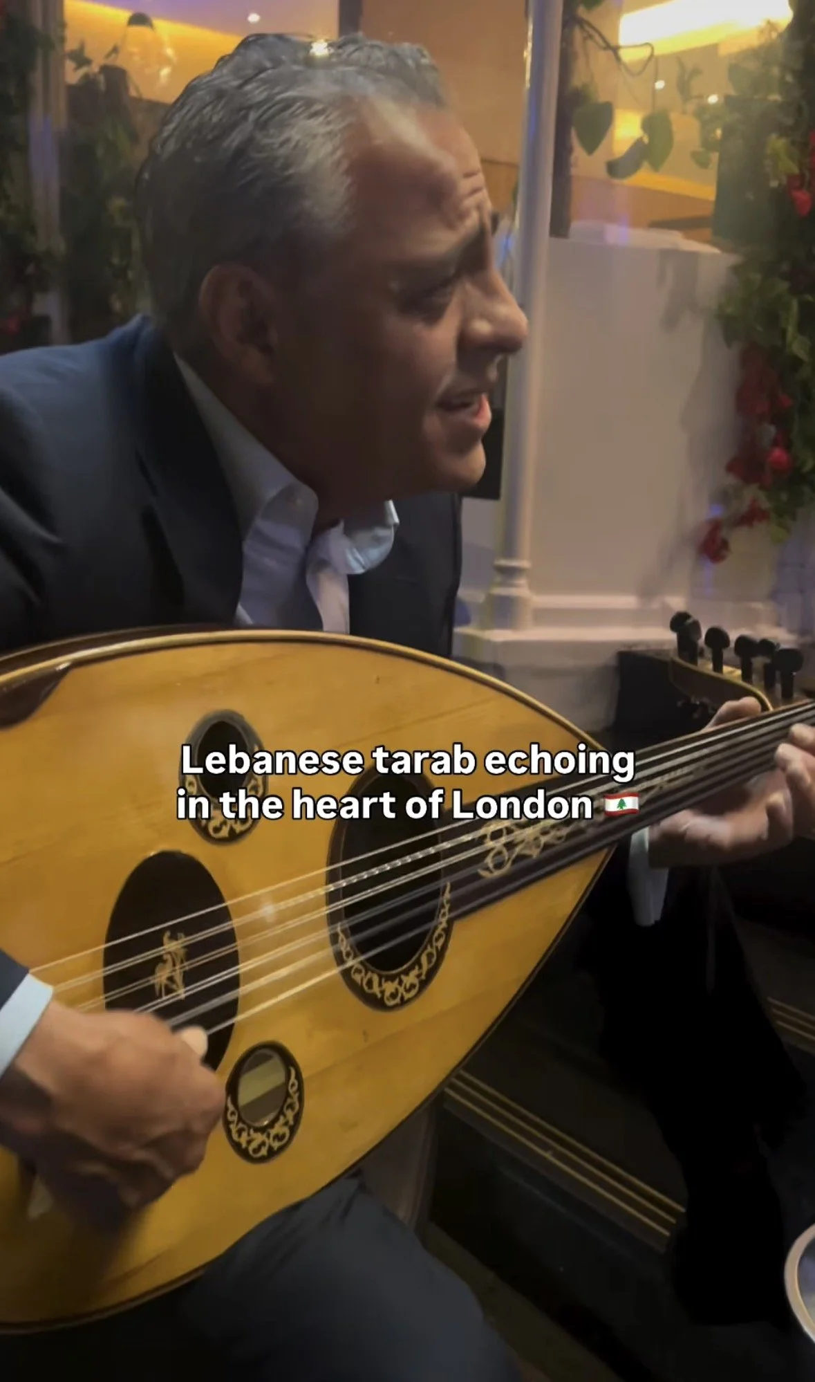 A man with gray hair playing a traditional Lebanese oud guitar in a cozy indoor setting with warm lighting and potted plants. The image includes the text 'Lebanese tarab echoing in the heart of London' with a Lebanon flag emoji.