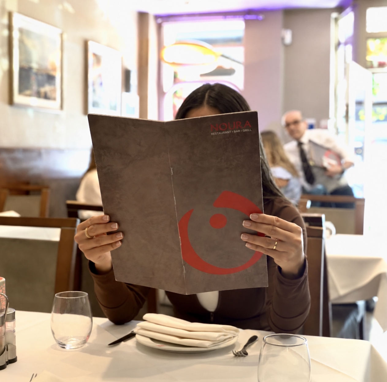 Woman sitting at a Noura table reading a menu, with plates, glasses, and utensils in front. The restaurant has a cozy ambiance with artwork on the wall and a man and woman in the background.