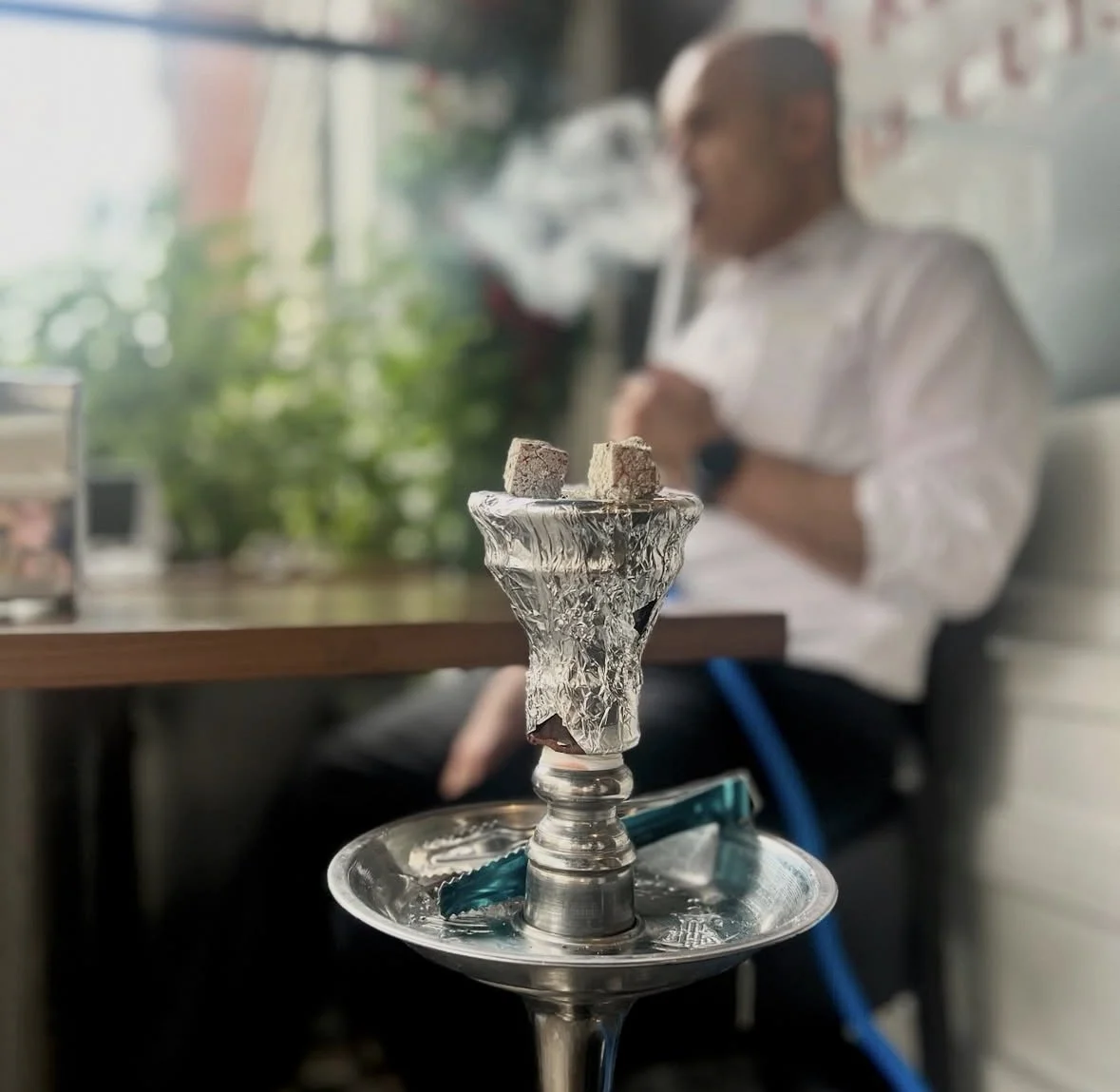 A hookah with a foil-wrapped tobacco bowl with three small cube-shaped flavor additives, in a cafe or lounge setting with a man in the background.