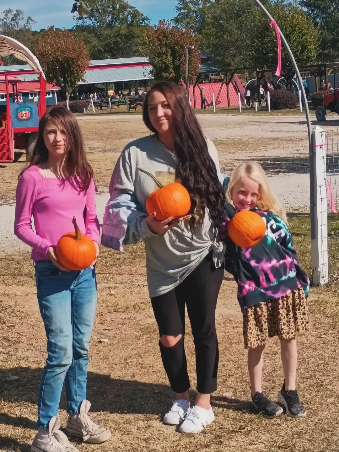 Three girls holding pumpkins outdoors on a fall day at a park or fairground.
