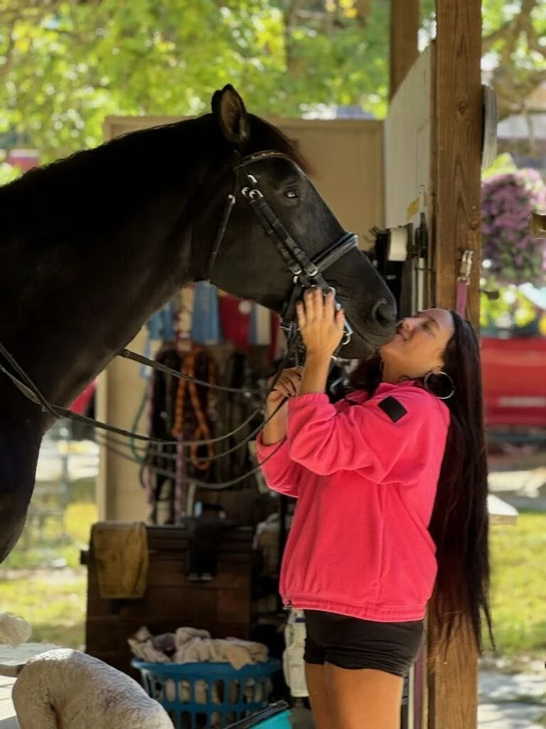 A woman in a pink jacket and black shorts standing close to a black horse, gently touching its face while leaning in for a kiss inside a barn with wooden beams and gear hanging in the background.