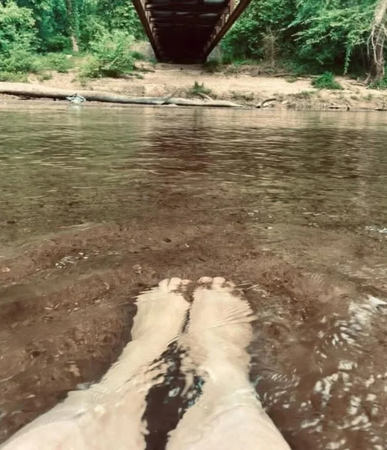 Photo taken from the perspective of someone standing in a river, showing their white legs and feet submerged in water. In the background, there's a bridge overhead and green trees along the riverbank.