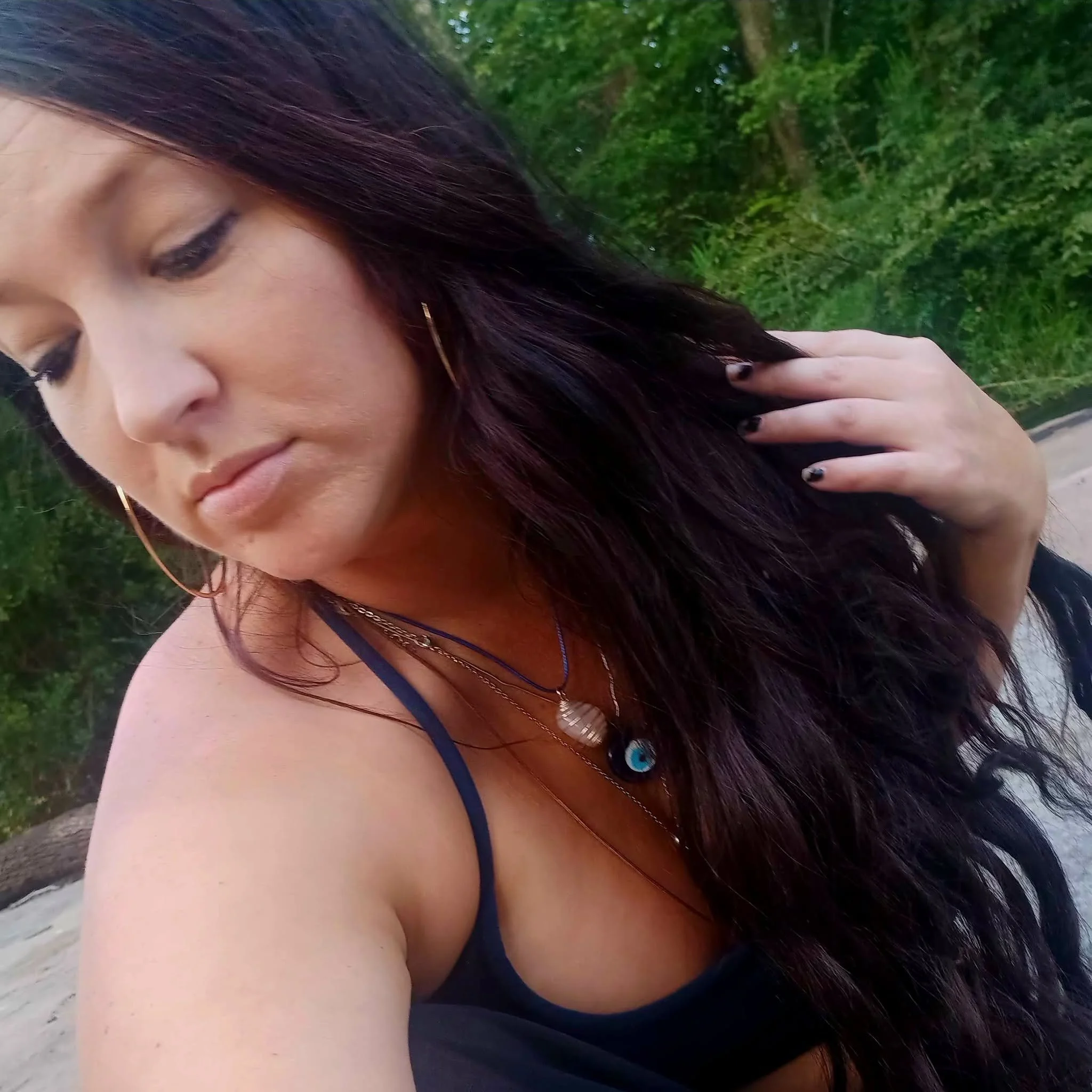 Close-up of a woman with long dark hair, wearing a black tank top with a blue strap, holding her hand near her face, outdoors with green trees in the background.