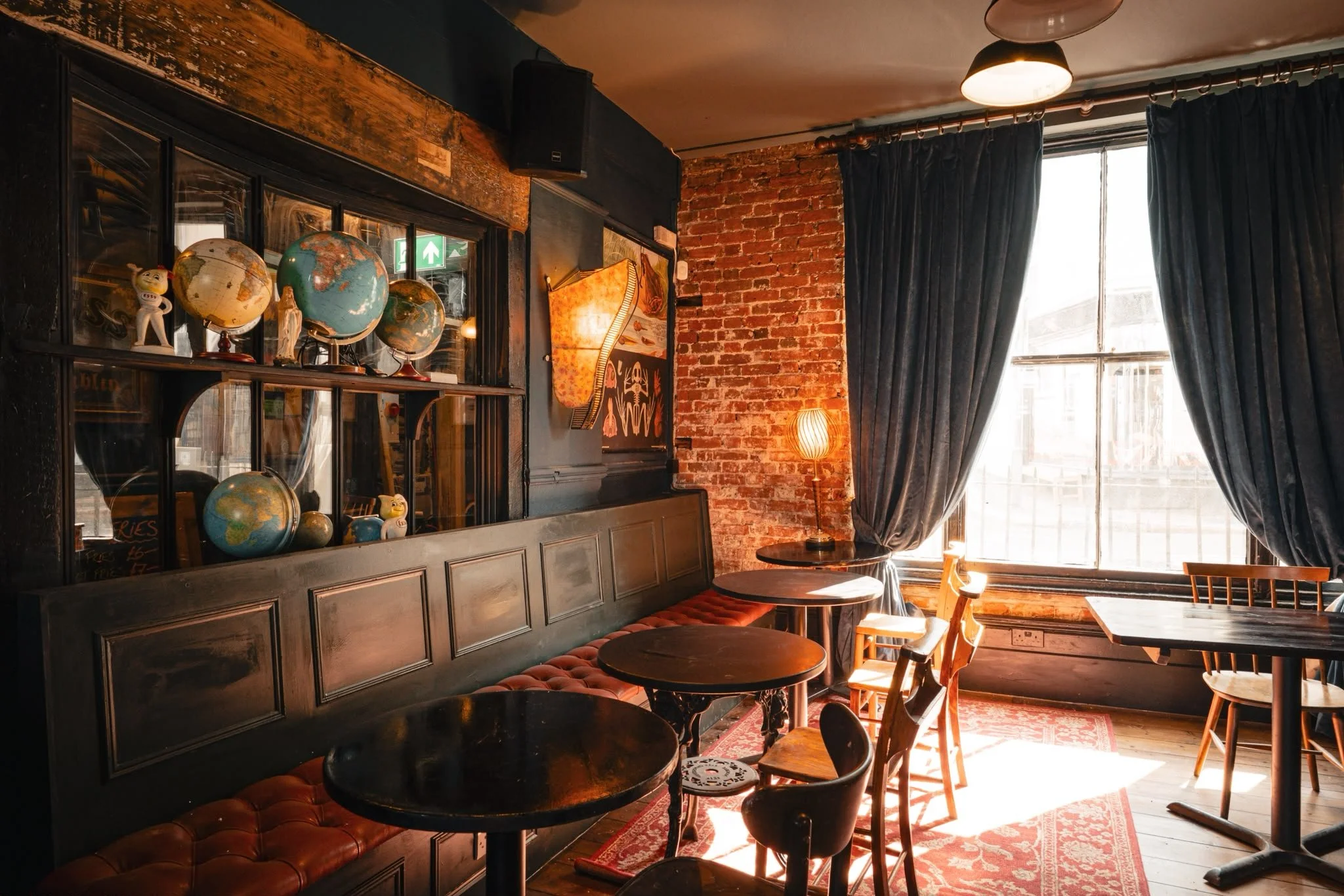 A cozy cafe with exposed brick walls, a large window with dark curtains, and a shelf with globes, small statues, and decorative items. There are round tables and chairs, warm lighting, and a patterned rug.