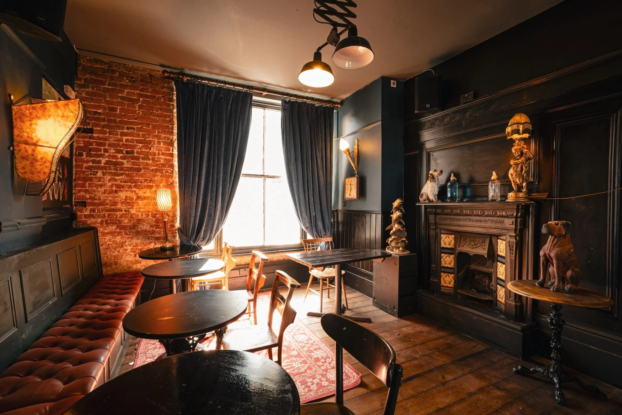 Interior of a cozy vintage-style cafe with dark walls, a brick accent wall, large window with dark curtains, wooden tables, and eclectic decor including a lamp, sculptures, and paintings.
