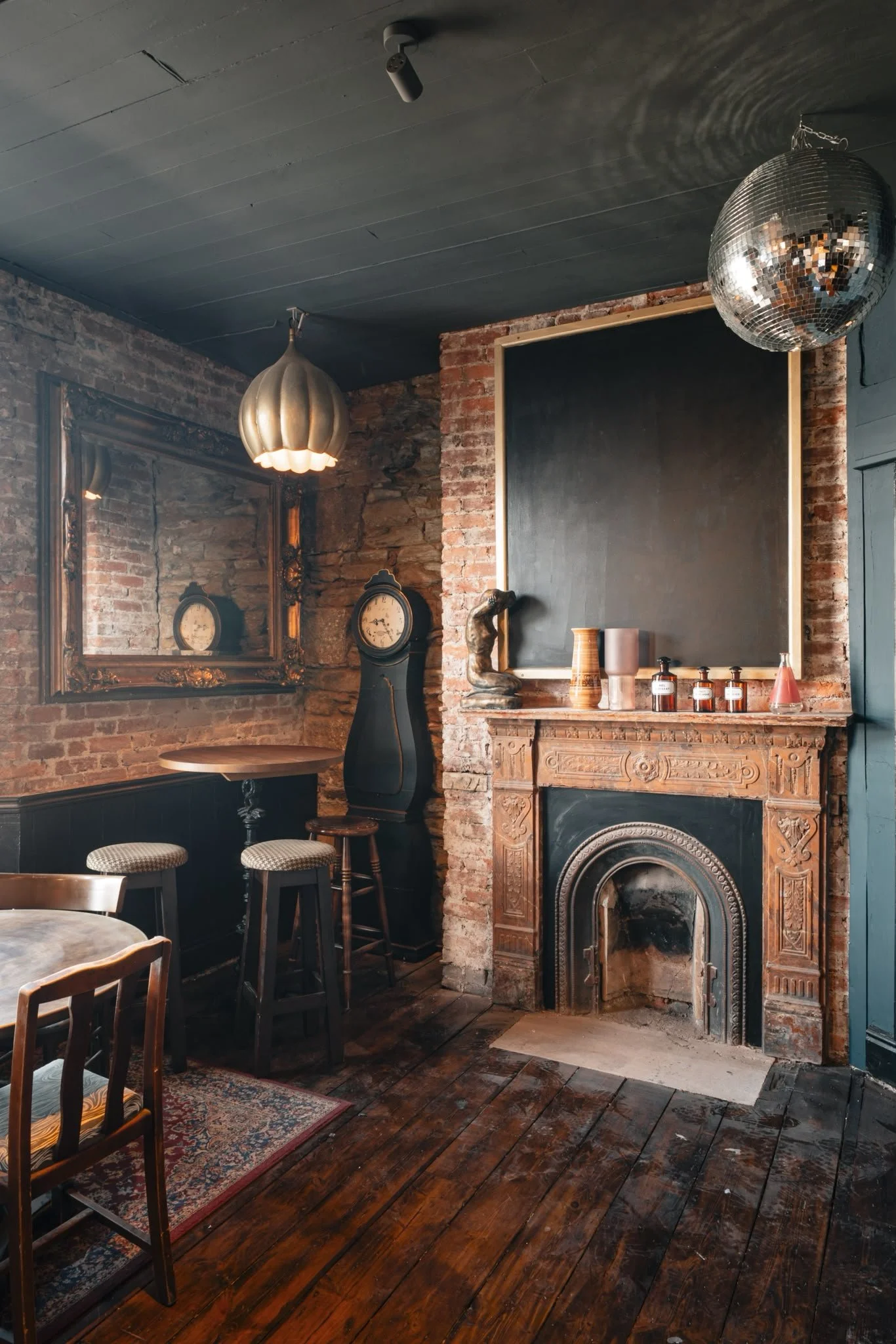 Interior of a cozy room with brick walls, a wooden floor, a vintage fireplace, small round stools and chairs, a mirror, a clock, and a large mirror or blackboard above the fireplace, with decorative vases and bottles on the mantel. A disco ball hangs from the ceiling.