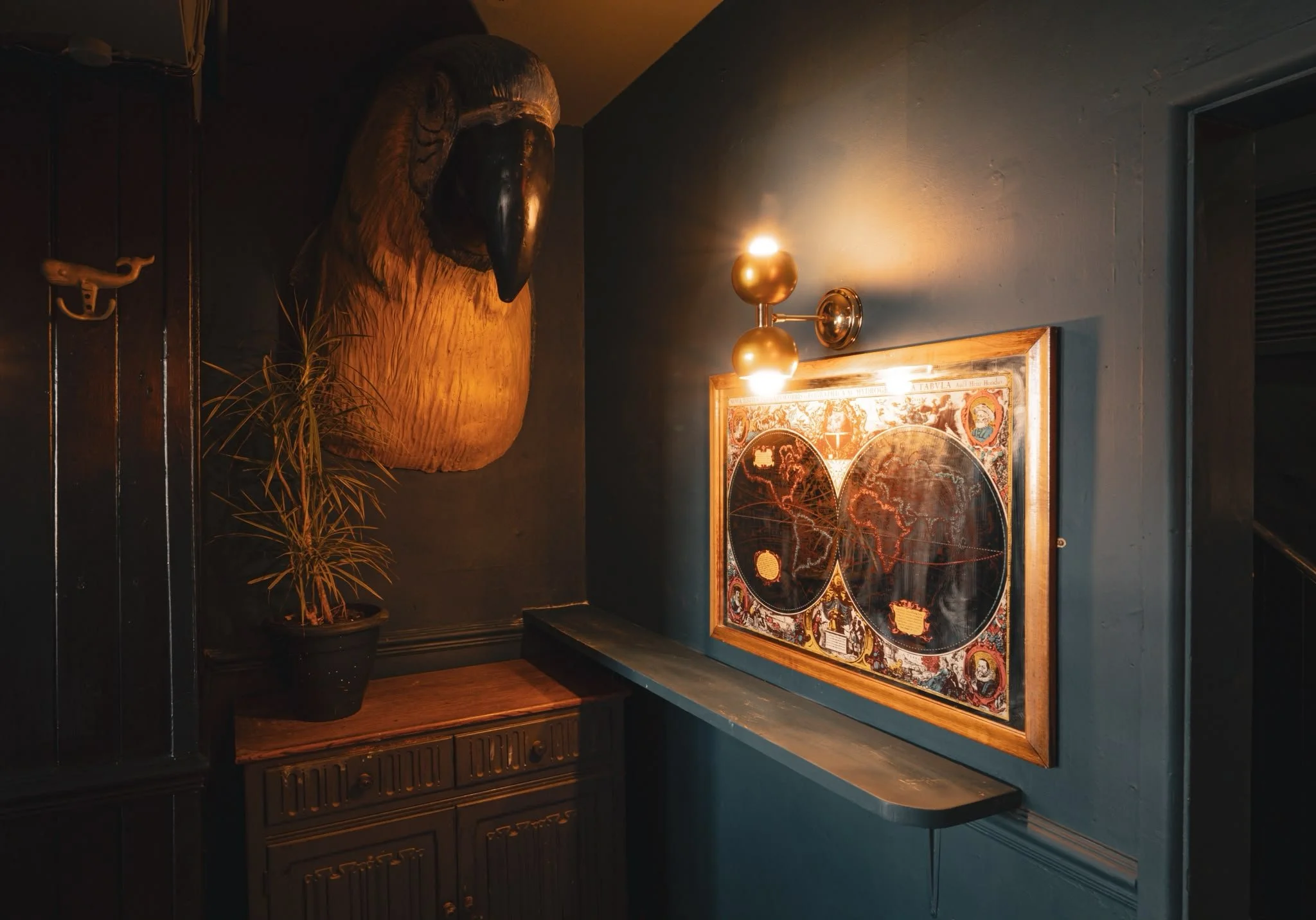 A dimly lit room featuring a mounted animal head, possibly a buffalo, above a framed vintage map. There is a small potted plant with dry grass nearby, a wall-mounted brass light fixture with two bulbs, and a black wooden panel with a decorative hook.