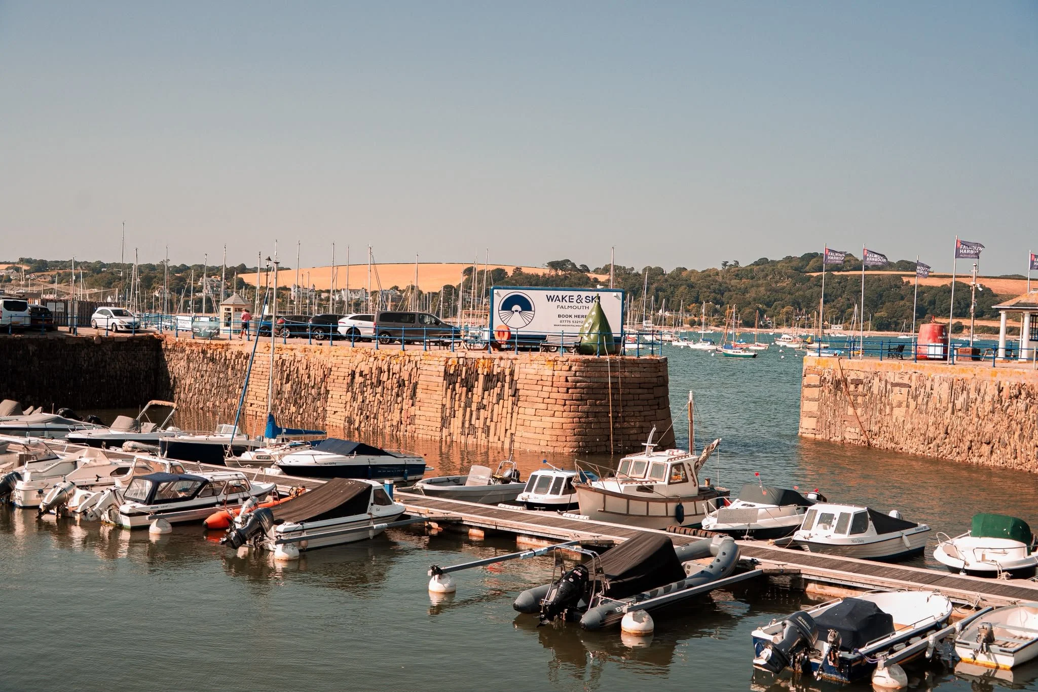 Marina with many boats docked, brick wall, parking lot with cars, hill with trees and flags, sign for Wake & Ski Falmouth.