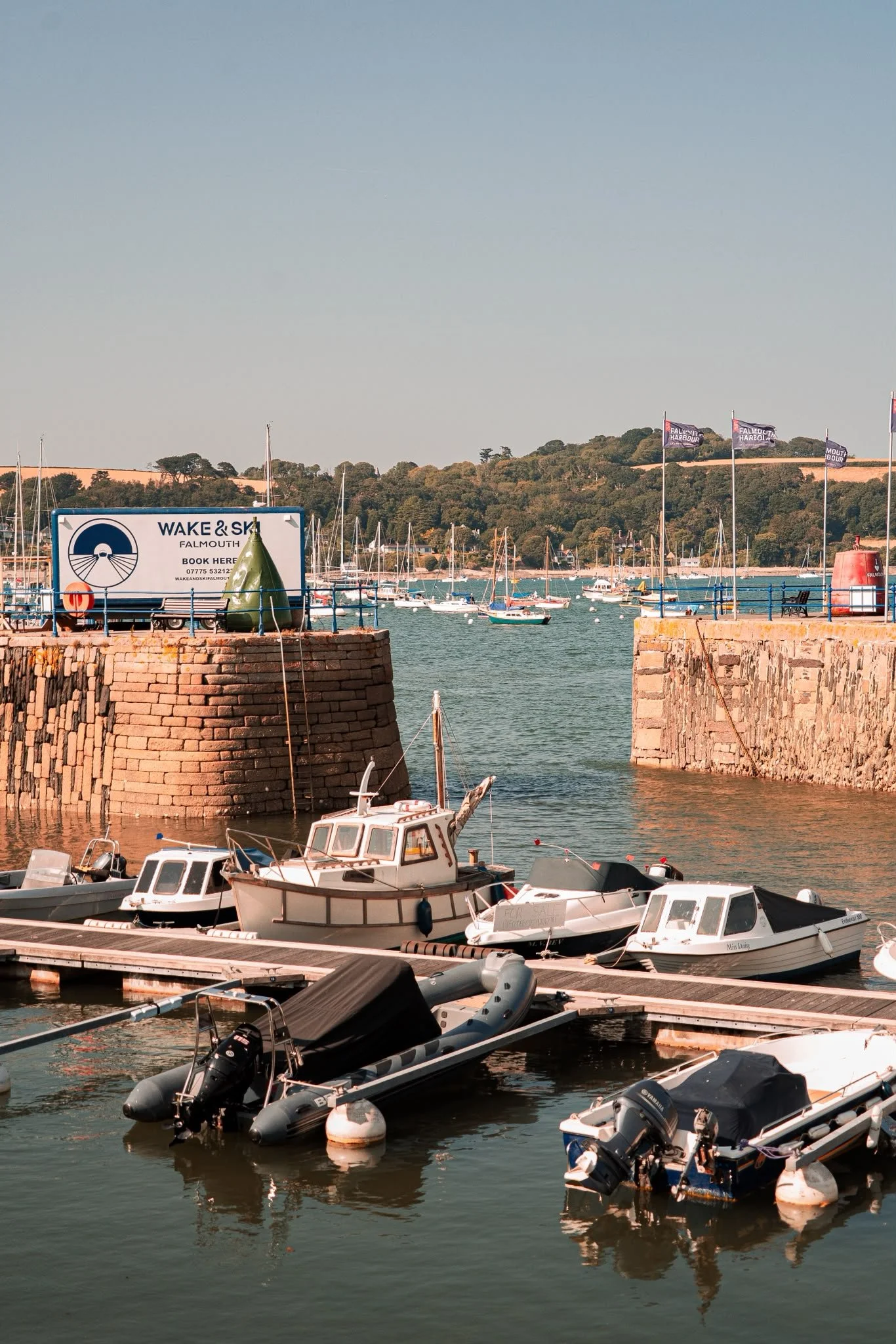 A marina with several boats docked along a pier, with a stone wall in the foreground and flags flying on a hill in the background.