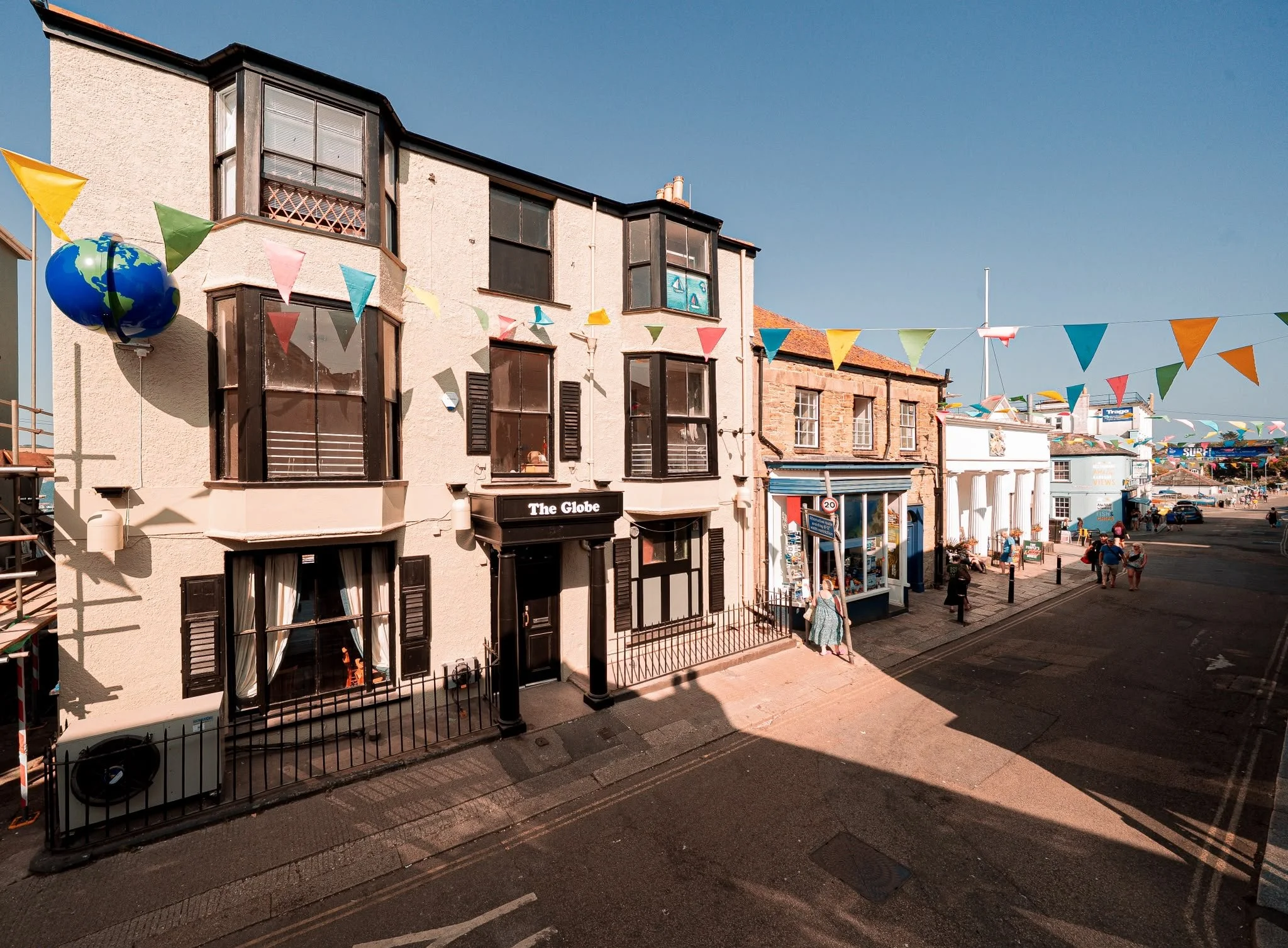 Street scene with buildings decorated with colorful bunting and a blue sky. People are walking along the sidewalk.