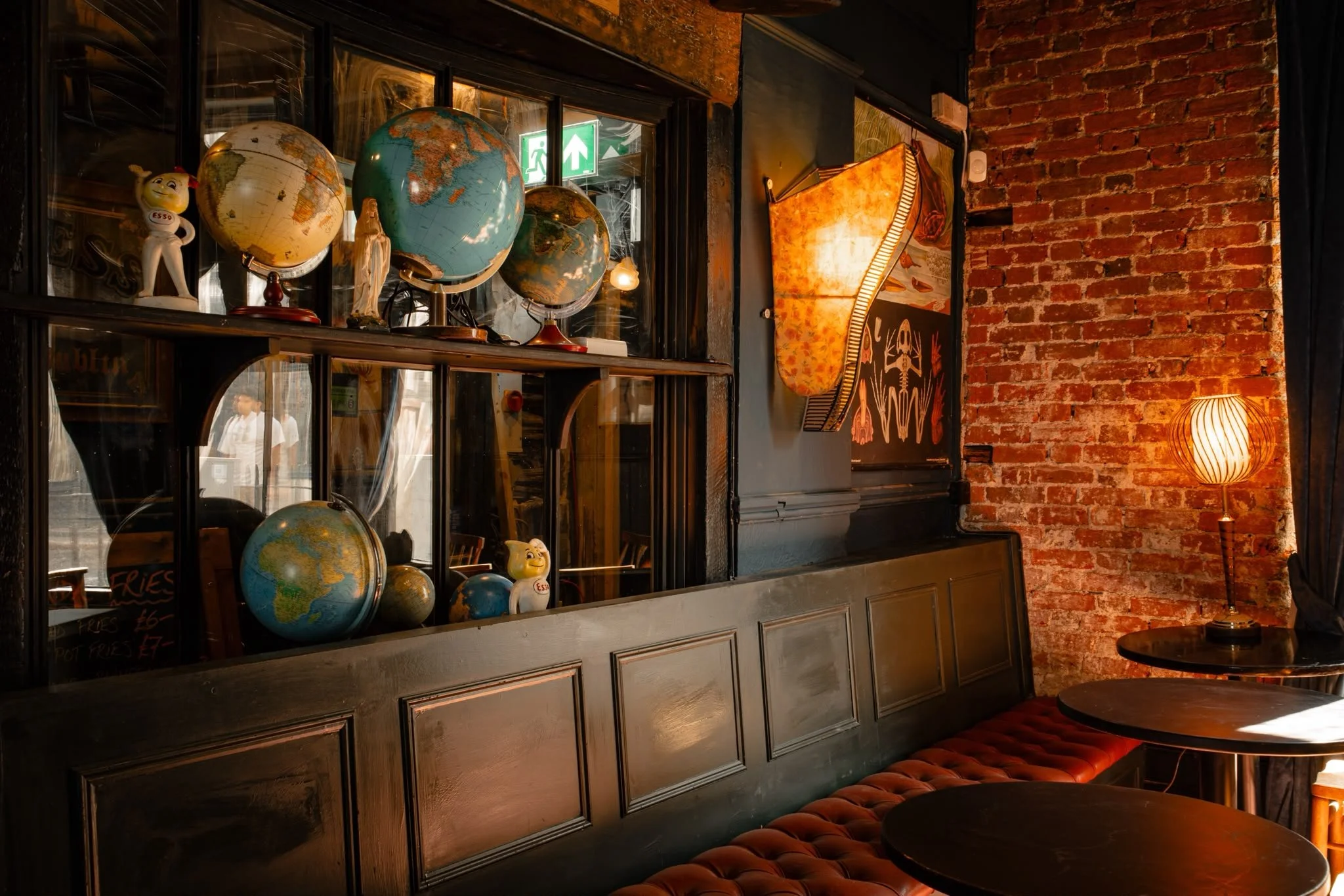 Interior of a cozy cafe with a brick wall, a shelving unit filled with globes and decorative items, a vintage-style lamp on the wall, and a small round table with a red cushioned bench.