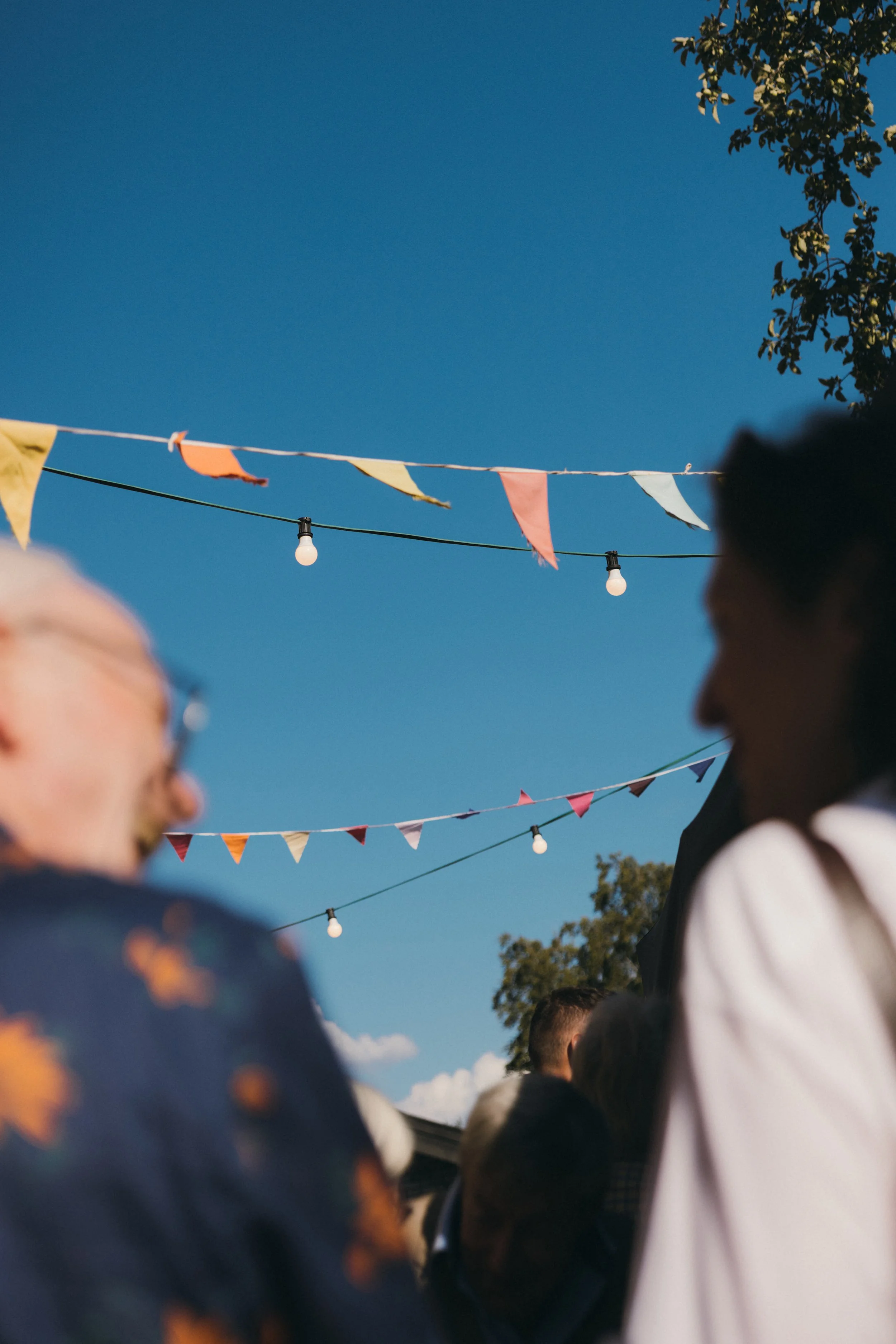 Menschen bei einem Fest im Freien, bunte Fähnchen und Lichterketten hängen in der Luft, blauer Himmel, Blick zwischen den Personen hindurch