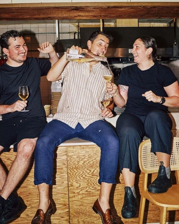 Drei Personen sitzen auf einer Bar, trinken Wein und lachen, während eine Person in der Mitte etwas eingießt.