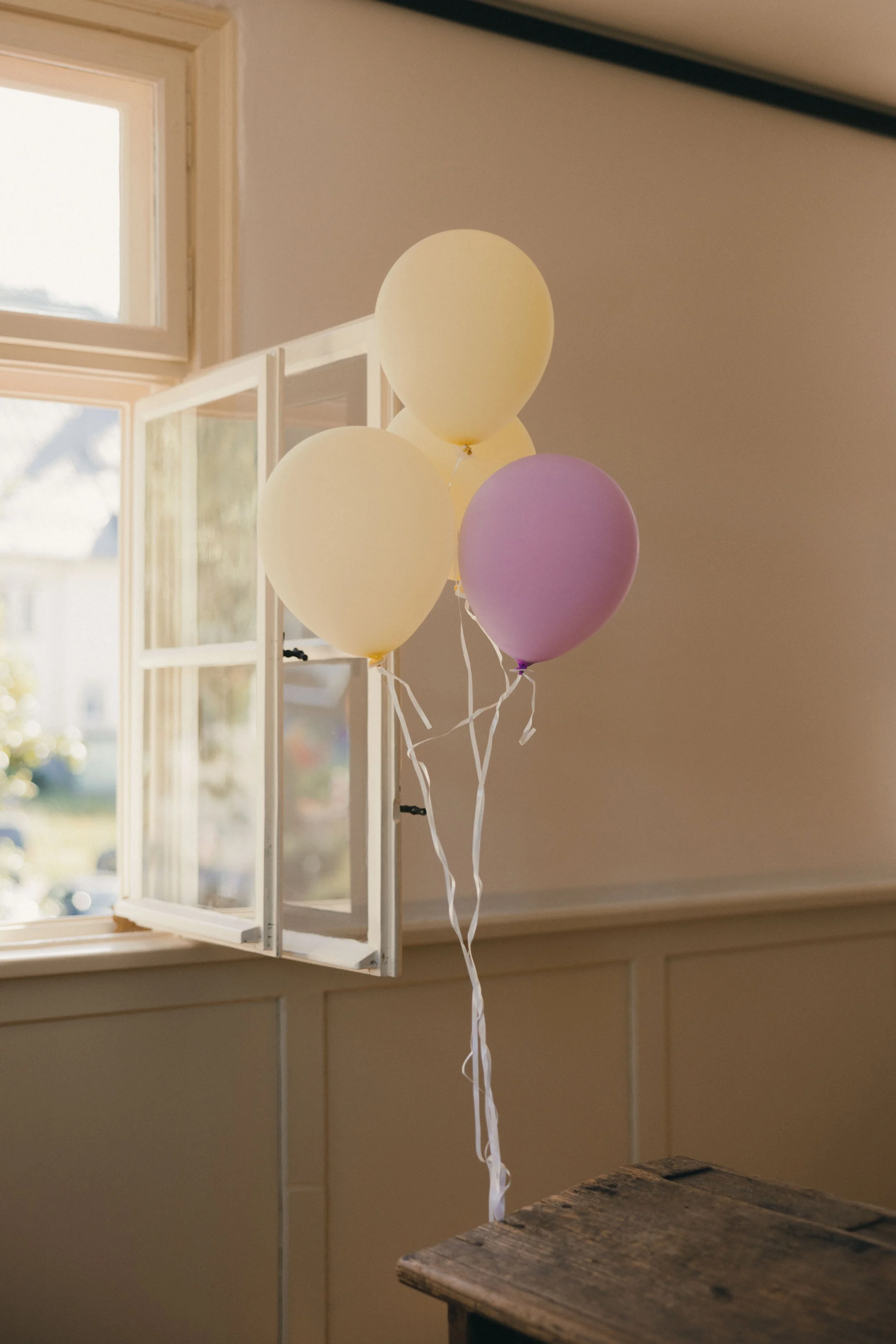 Bunte Ballons (gelb, lila, beige), die an einem Fenster hängen, im Innenraum, mit Sonnenlicht, das durch das Fenster scheint.