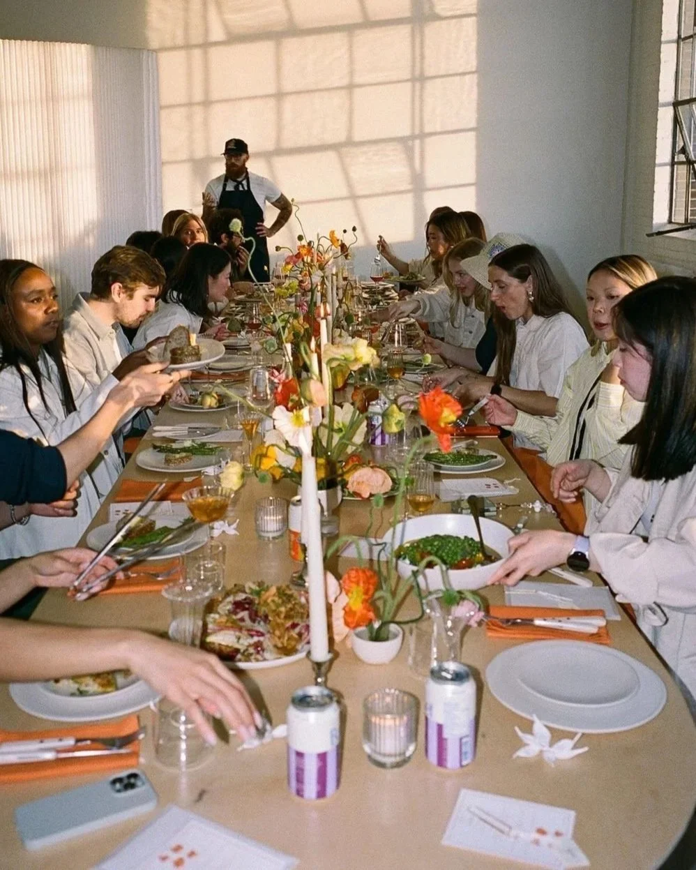 Menschen sitzen an einem lang gedeckten Esstisch mit Blumen, Kerzen und Geschirr bei einer festlichen Mahlzeit.
