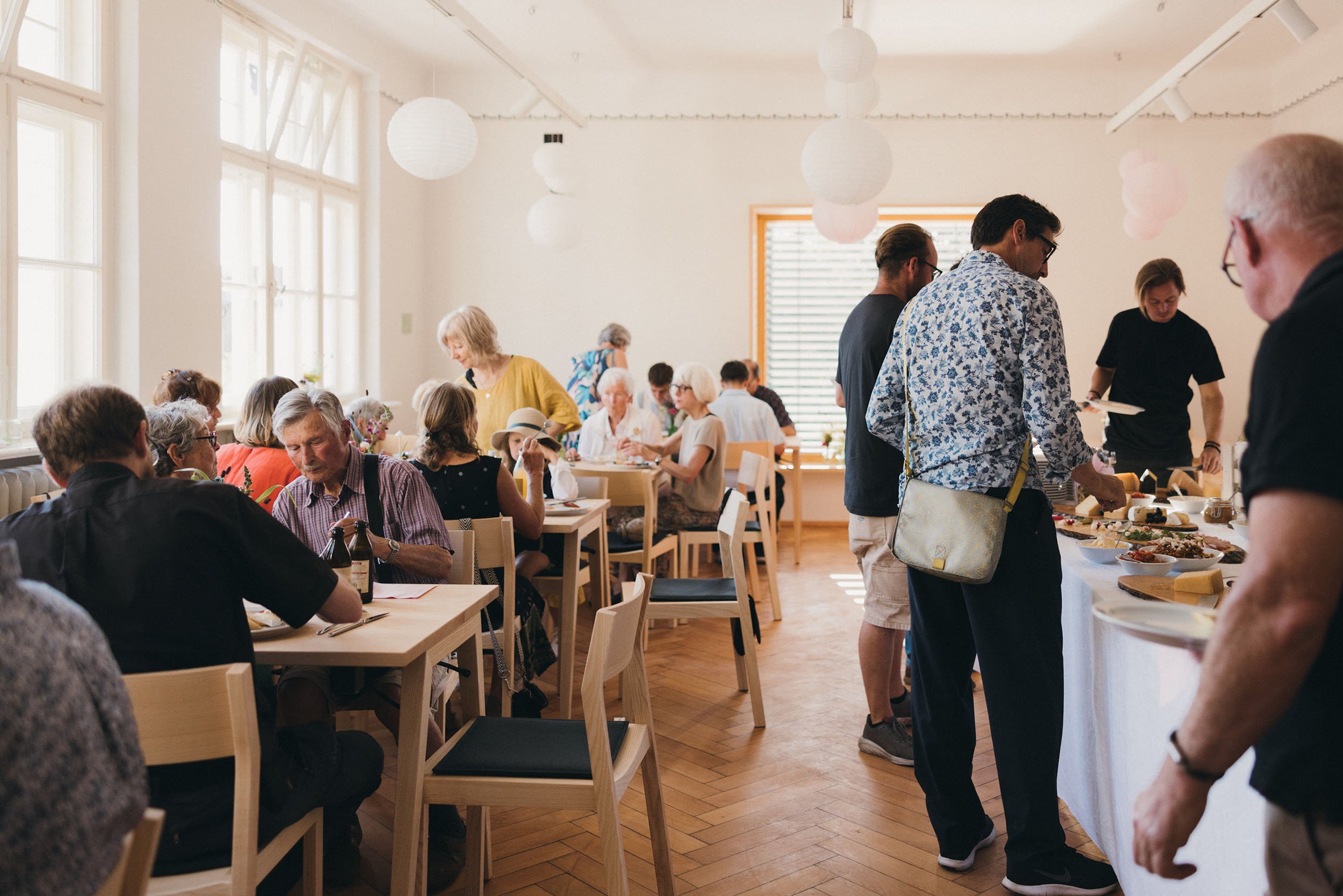 Menschen versammeln sich in einem hellen, gemütlichen Raum bei einem Buffet mit Speisen. Es herrscht eine freundliche Atmosphäre, viele sitzen oder stehen und unterhalten sich.